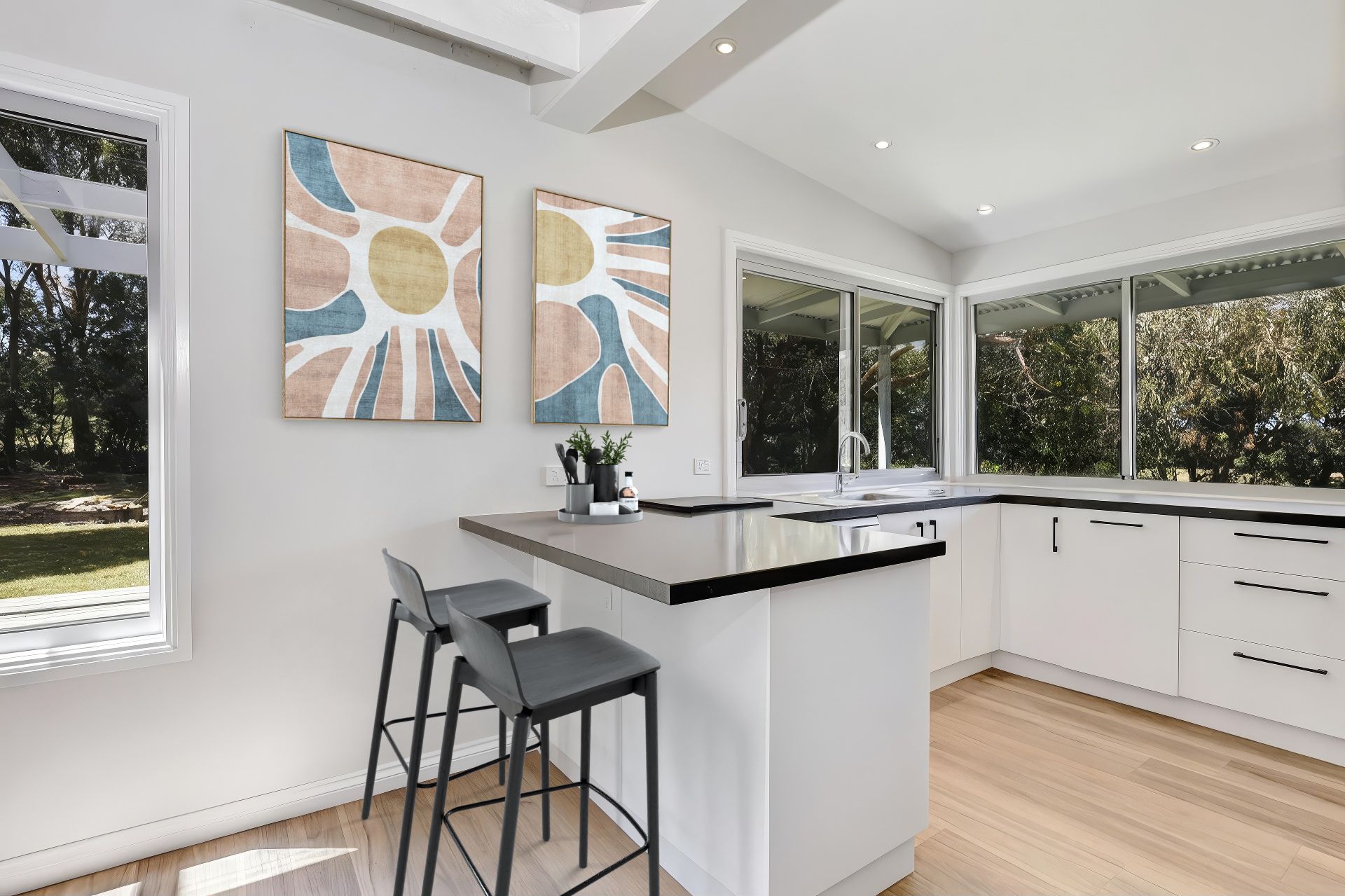 Modern white kitchen with a small island and bar stools, two art pieces, and large windows overlooking trees.