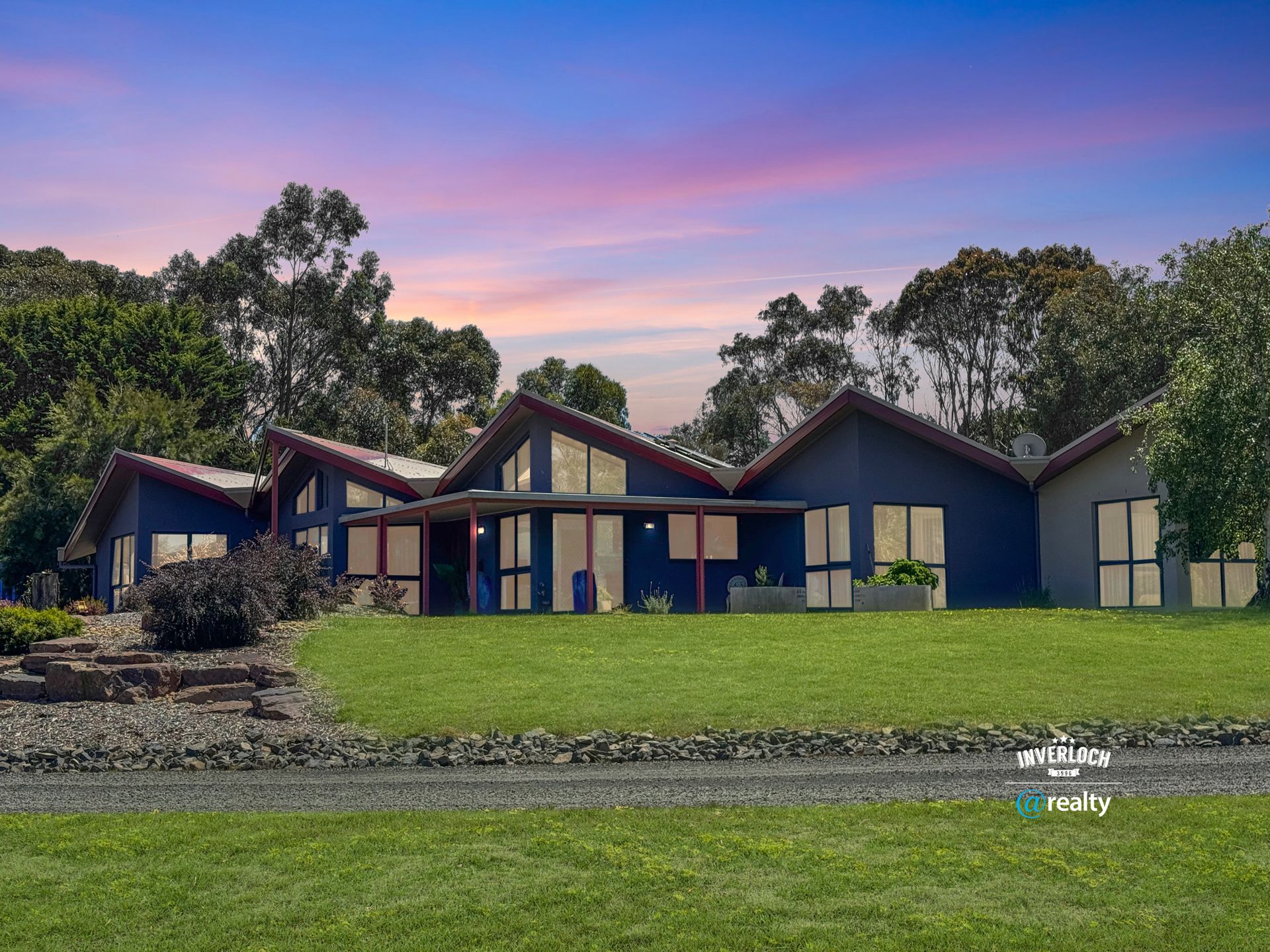 Blue house with multiple gabled sections on green lawn under a sunset sky, trees in the background.
