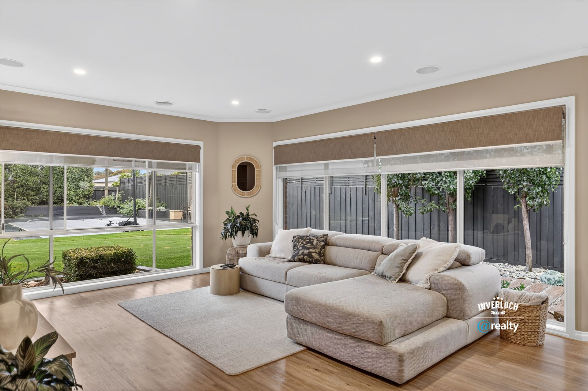 A bright, neutral-toned living room features a beige sectional sofa on a light rug, positioned near large corner windows.