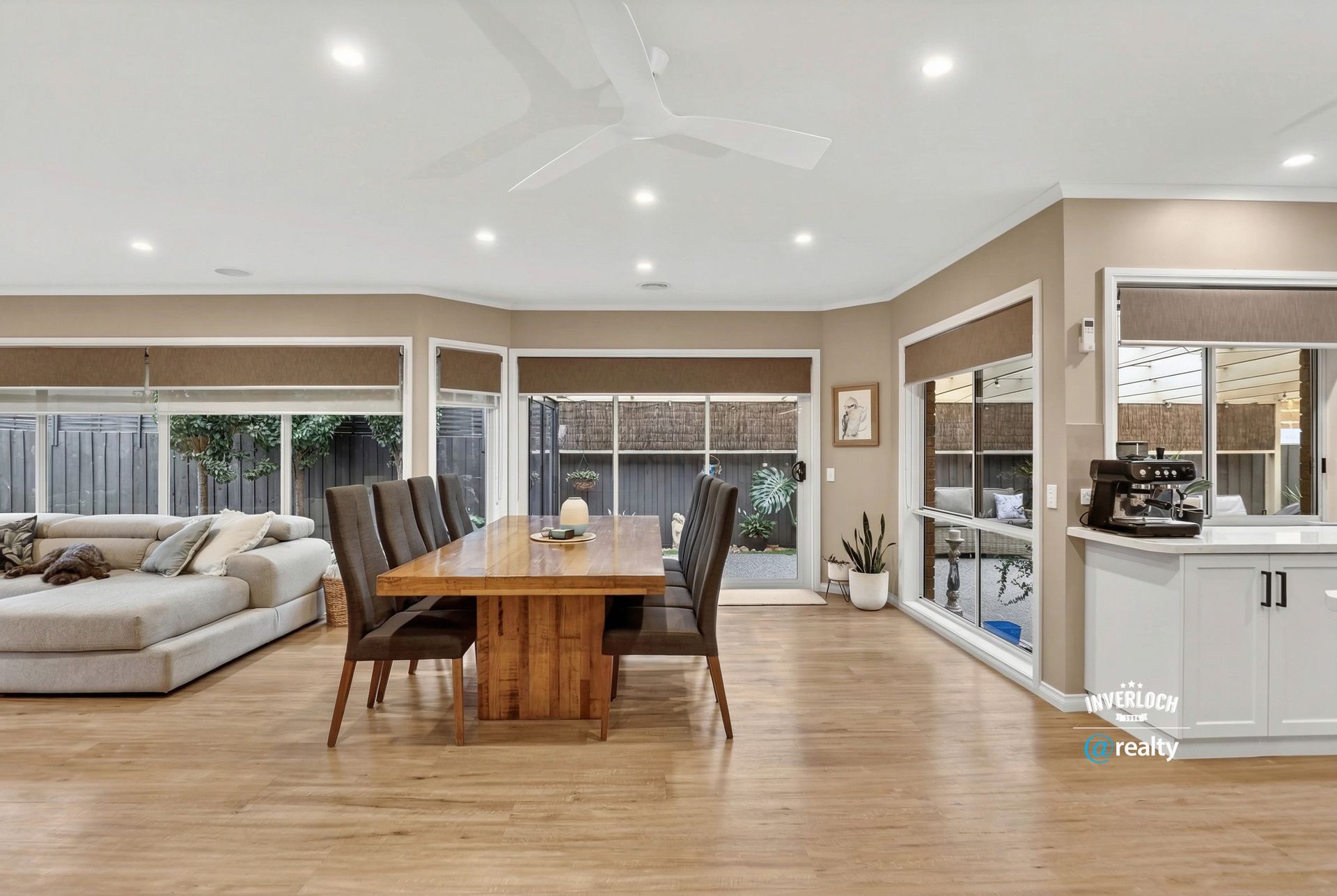 An open-plan living and dining area with a wooden table, grey sectional sofa, and kitchen counter on light wood floors.