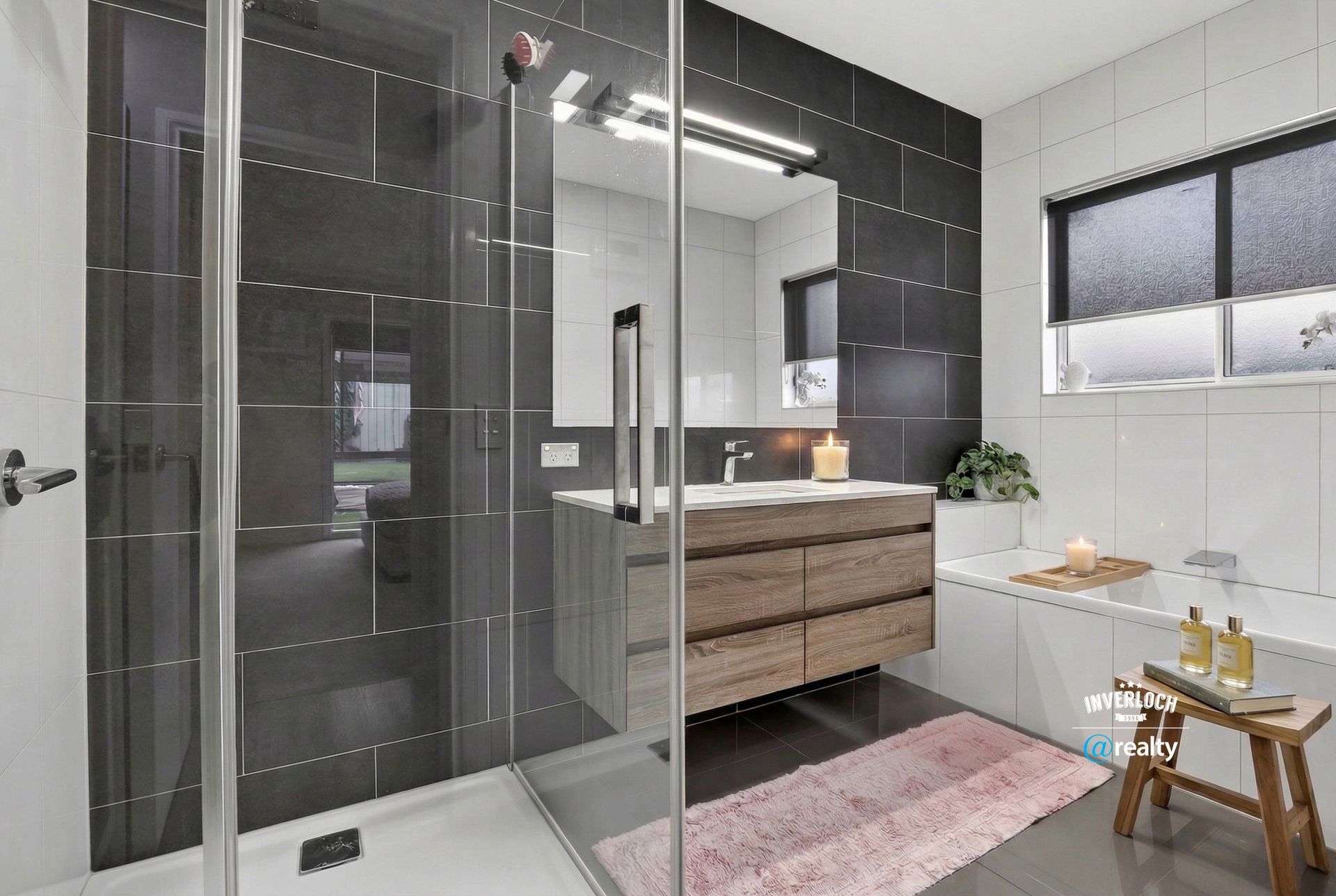 A modern bathroom with a glass-enclosed shower, a wood-finish vanity, a bathtub, and a pink rug on a grey tiled floor.