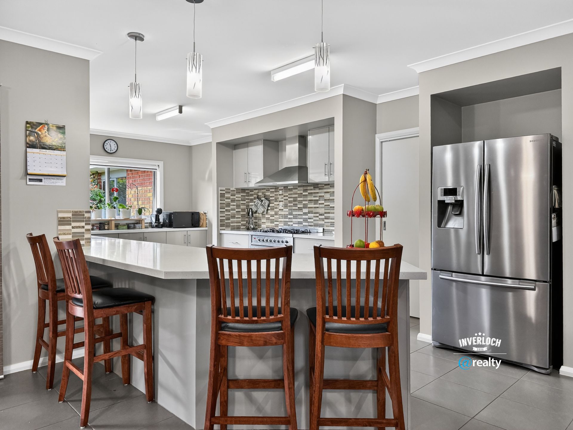 A bright kitchen with a granite island, dark wooden stools, stainless steel refrigerator, and modern pendant lighting.