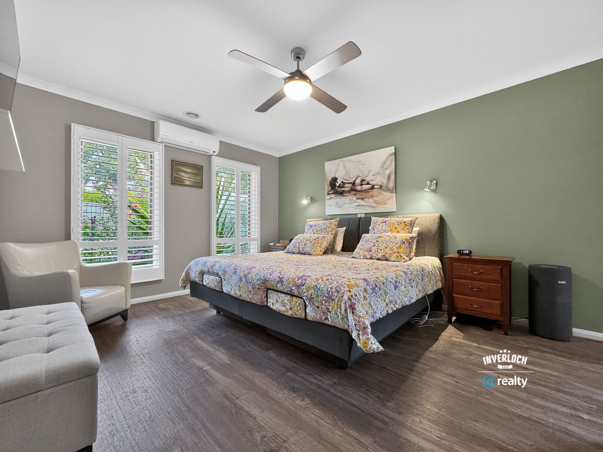 A bedroom featuring a large bed with floral bedding, grey walls, a sage green accent wall, wooden furniture, and a fan.