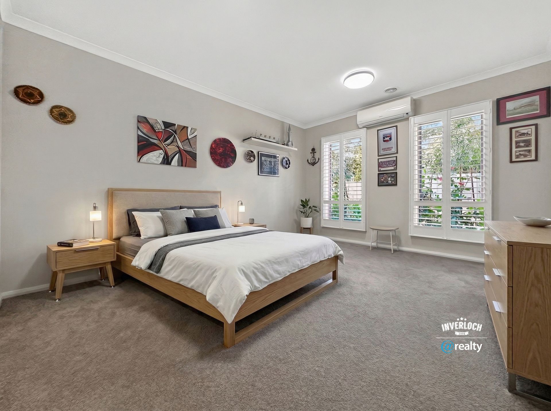 A spacious, neutral-toned bedroom with a large bed, beige walls, wall art, two tall windows, and a wooden dresser.