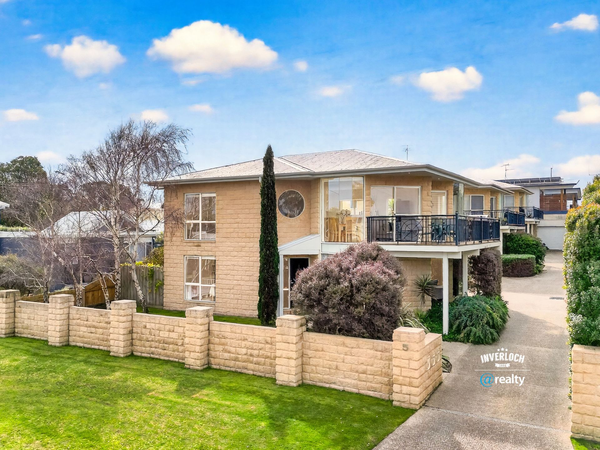 Two-story brick townhouses with balconies, surrounded by a stone wall and green lawn, under a blue sky.