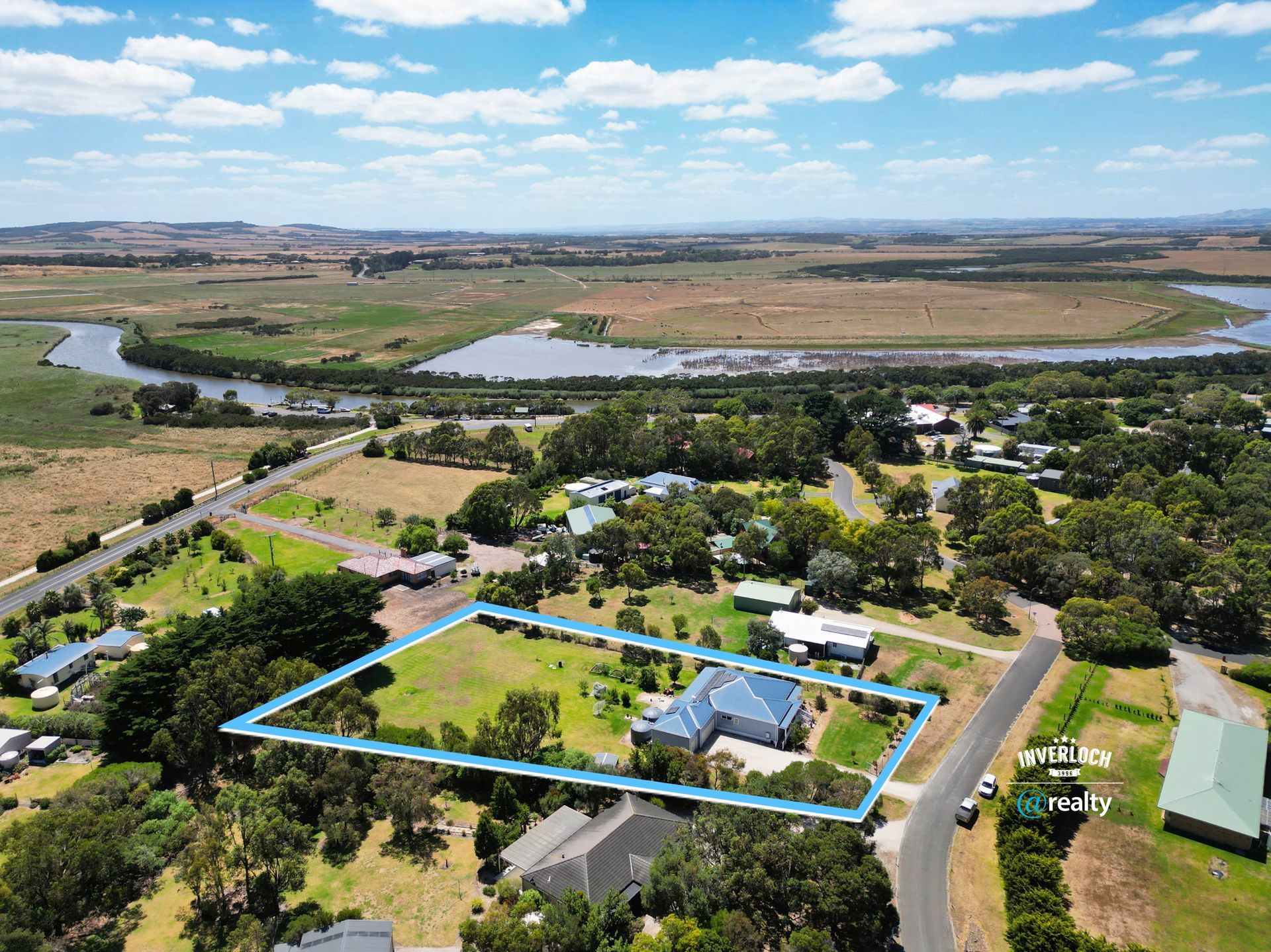Aerial view of a house with a blue roof, outlined in blue, surrounded by green grass and trees, near a river and field.