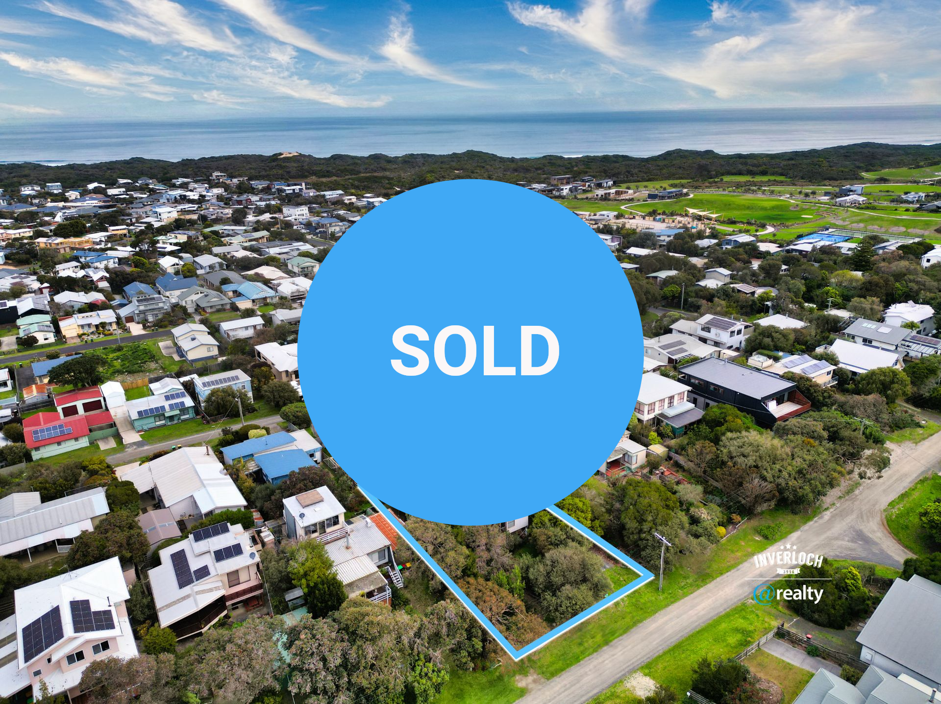 Aerial view of a coastal neighborhood with houses, trees, a road, and the ocean in the distance under a blue sky.
