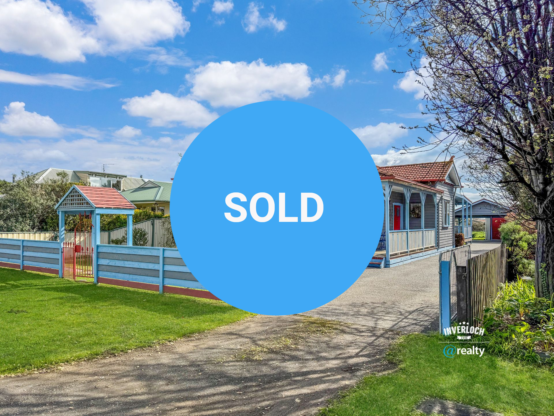 Blue cottage with red roof and porch, blue fence, driveway. Green grass, blue sky.