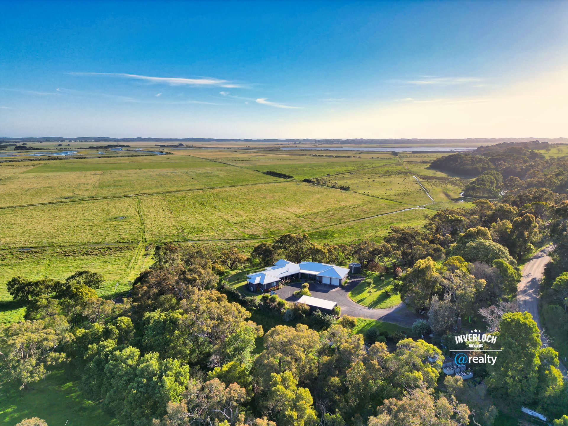 House surrounded by trees, overlooking a field at sunset. Long driveway.