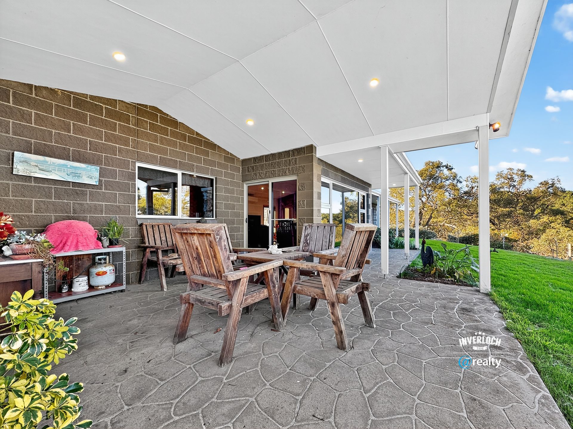 Patio with wooden furniture under a white covered roof, with a view of trees and a green lawn.