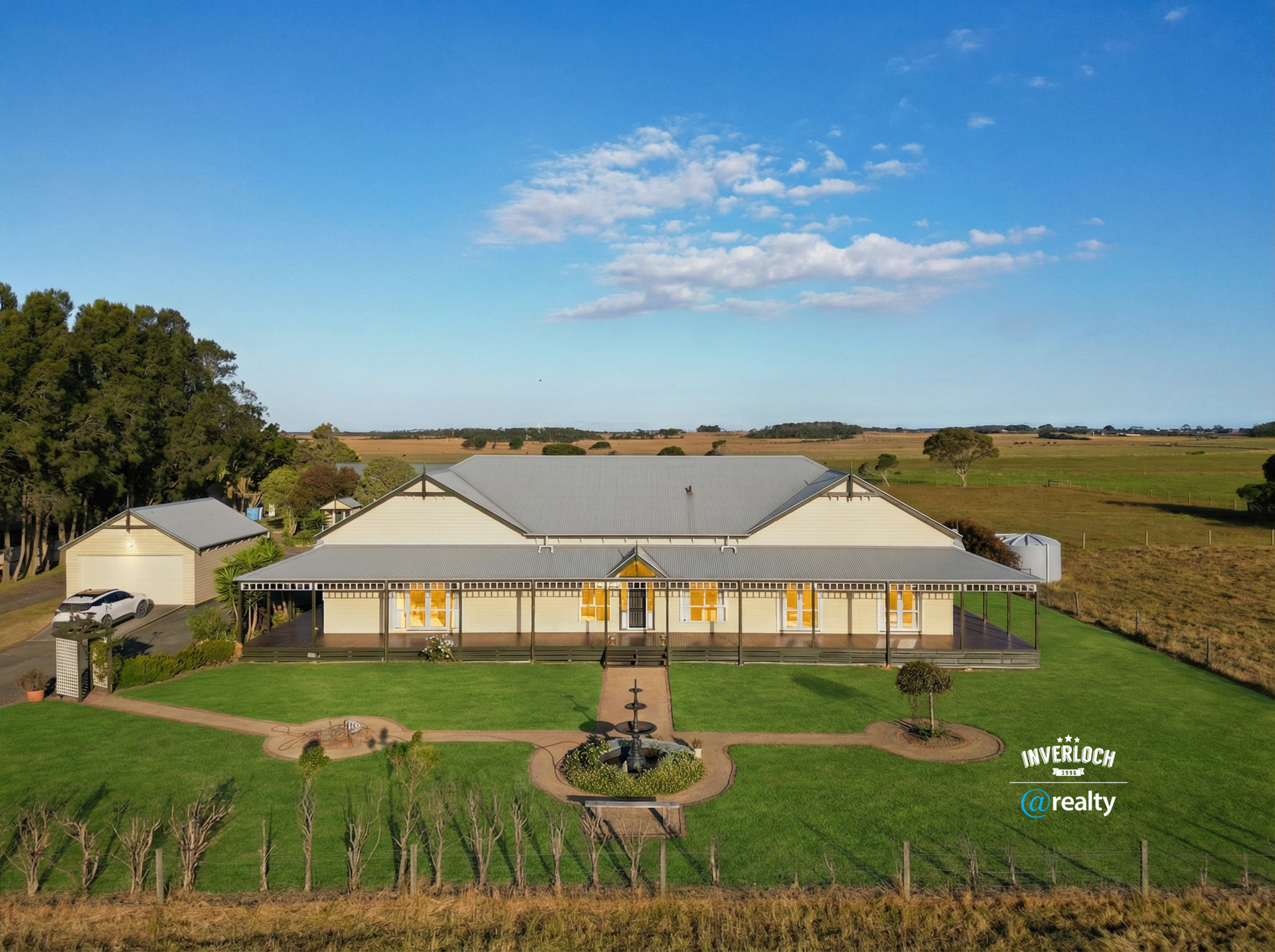 Large, light-colored house with a wraparound porch and fountain, on a grassy lawn, in a rural setting.