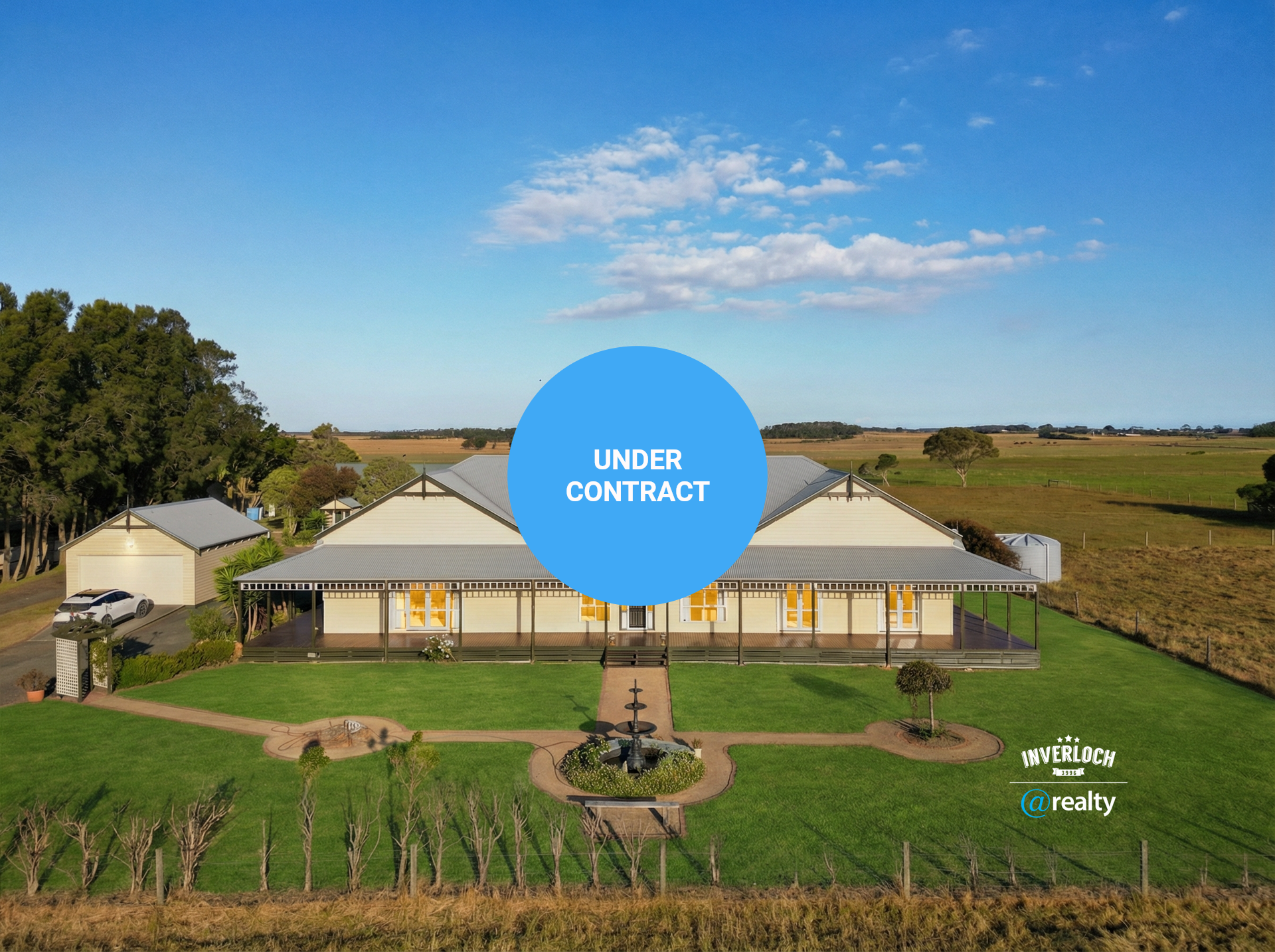Large, light-colored house with a wraparound porch in a grassy field on a sunny day. A fountain and vineyard are in front.