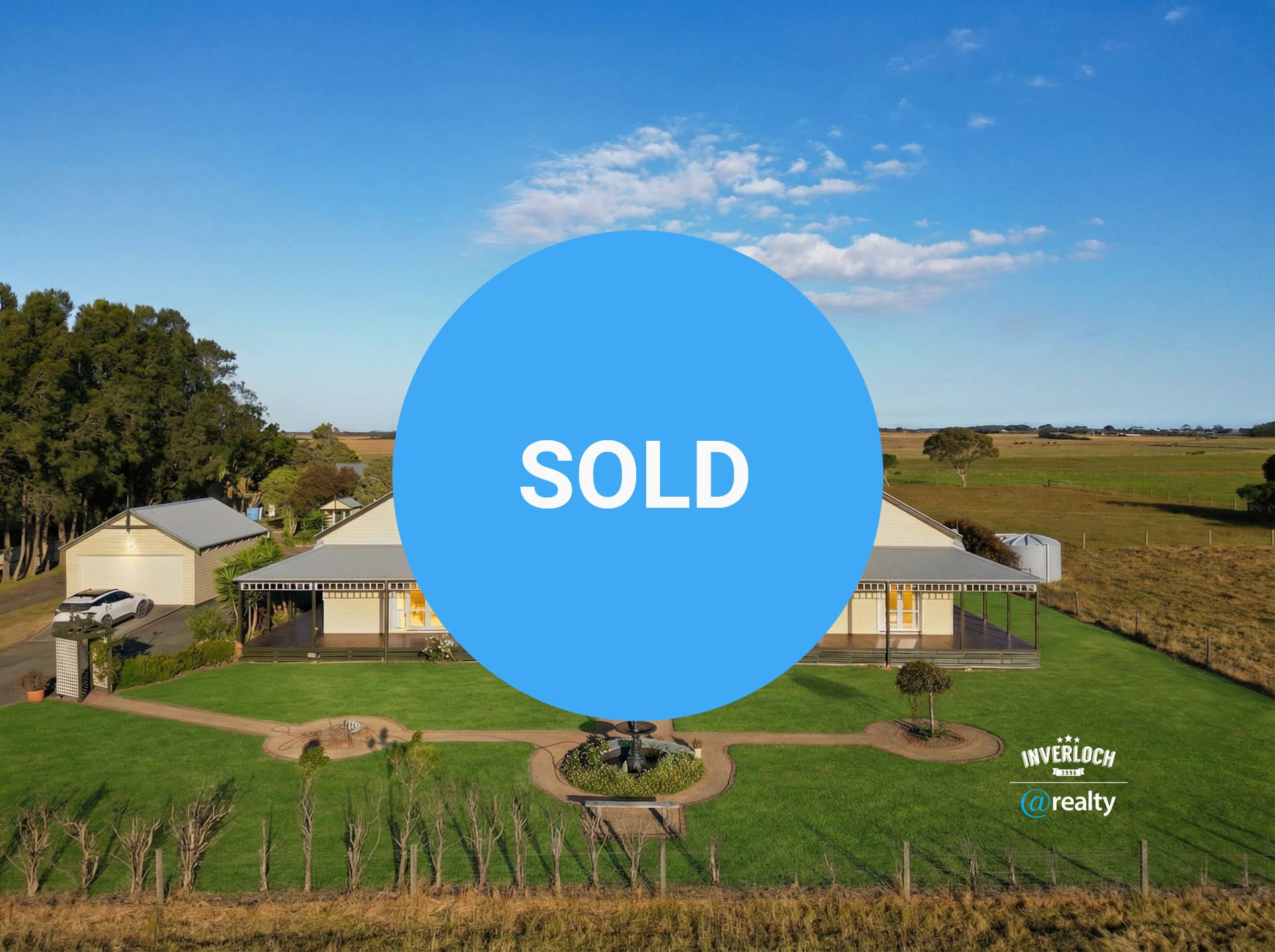 Large, light-colored house with a wraparound porch and fountain, on a grassy lawn, in a rural setting.