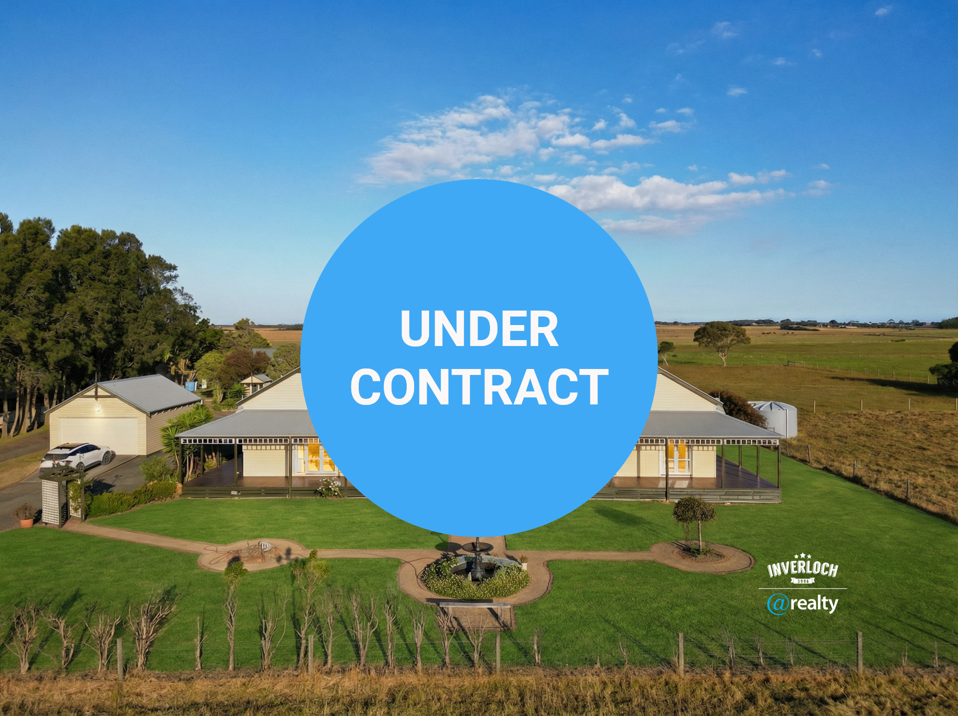Large beige house with gray roof, surrounded by green lawn and fields under a blue sky.