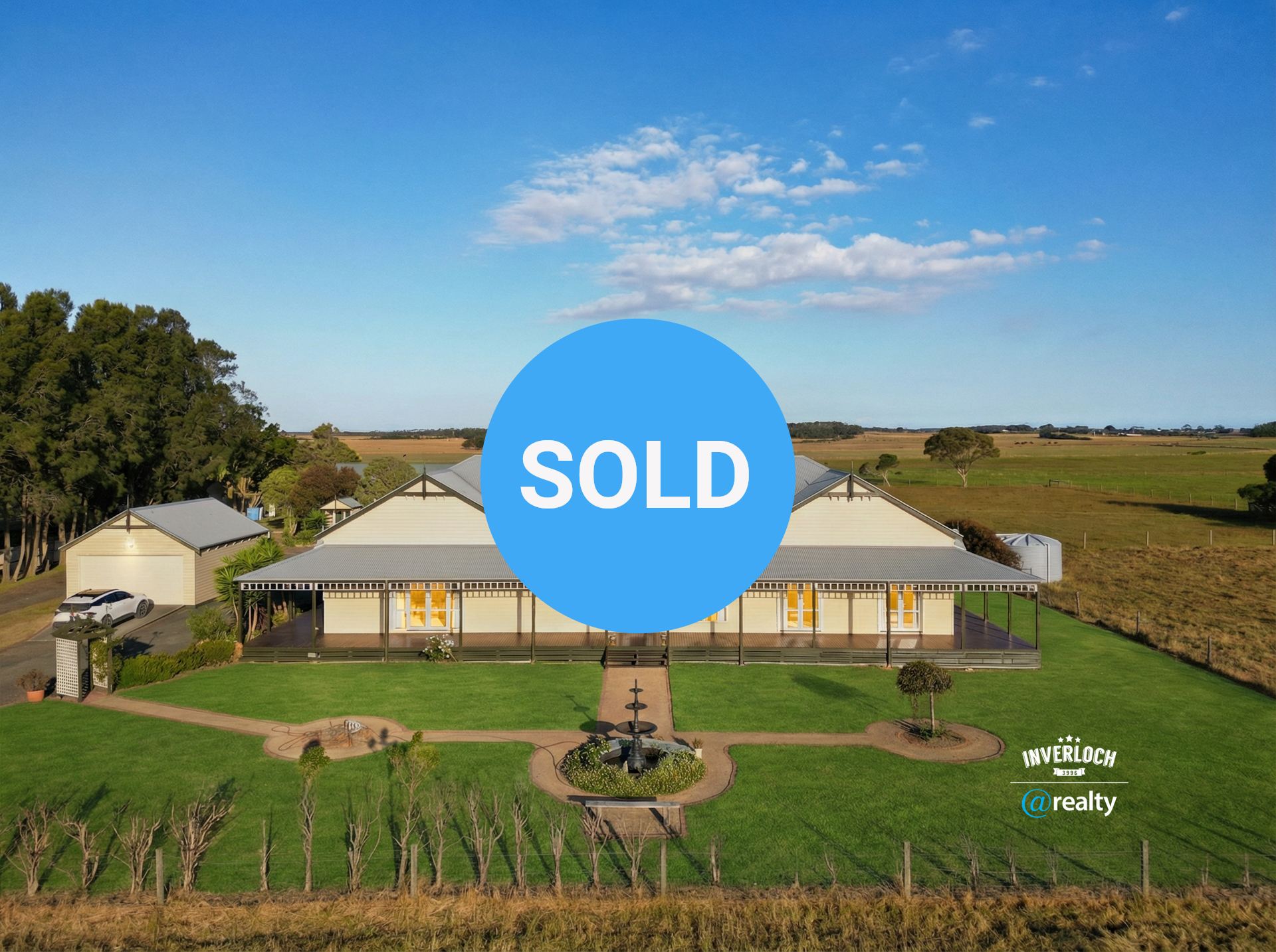 Large, light-colored house with a wraparound porch in a grassy field on a sunny day. A fountain and vineyard are in front.