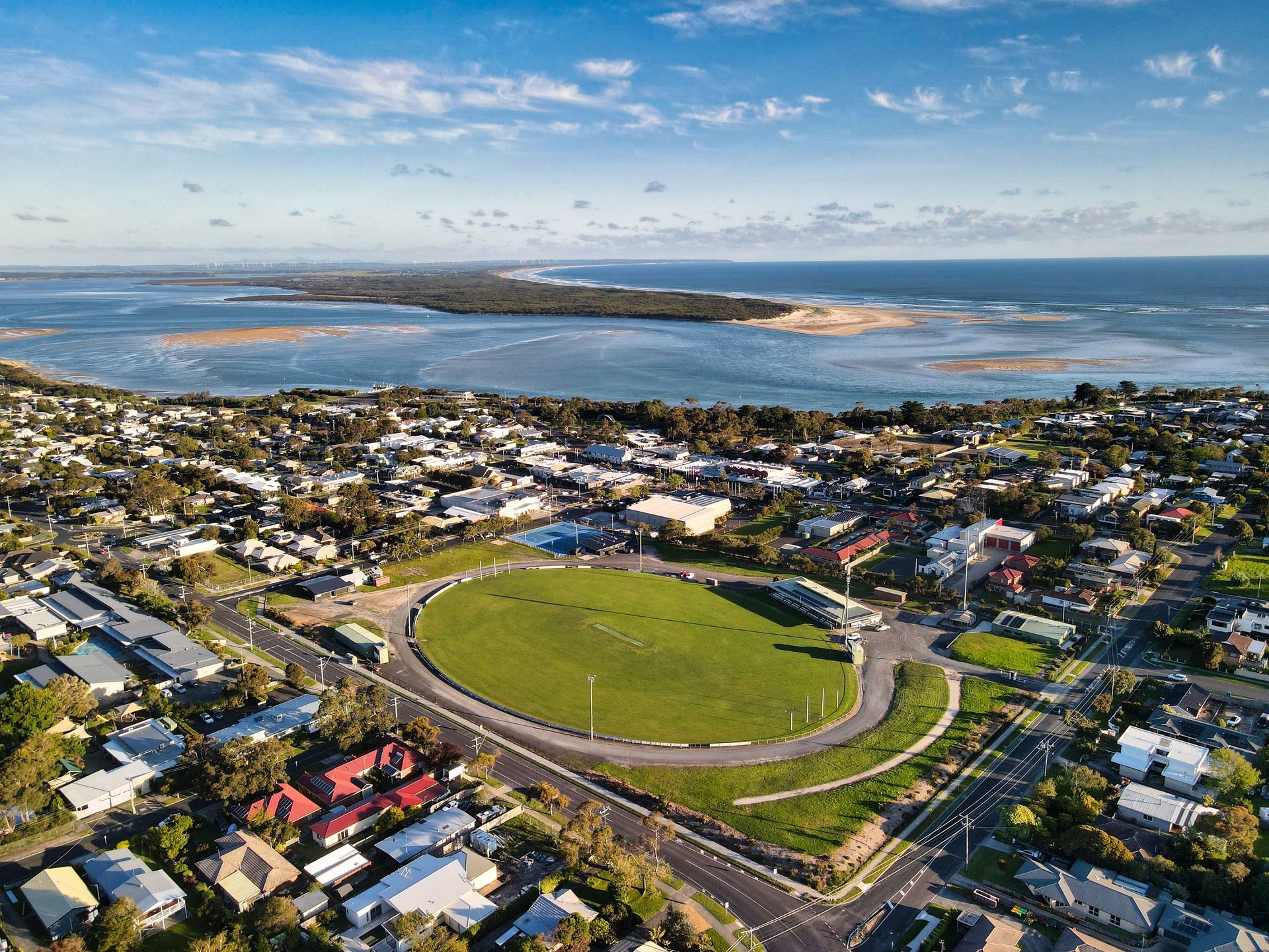 Aerial view of a coastal town with a large oval-shaped green field, buildings, and ocean in the background.