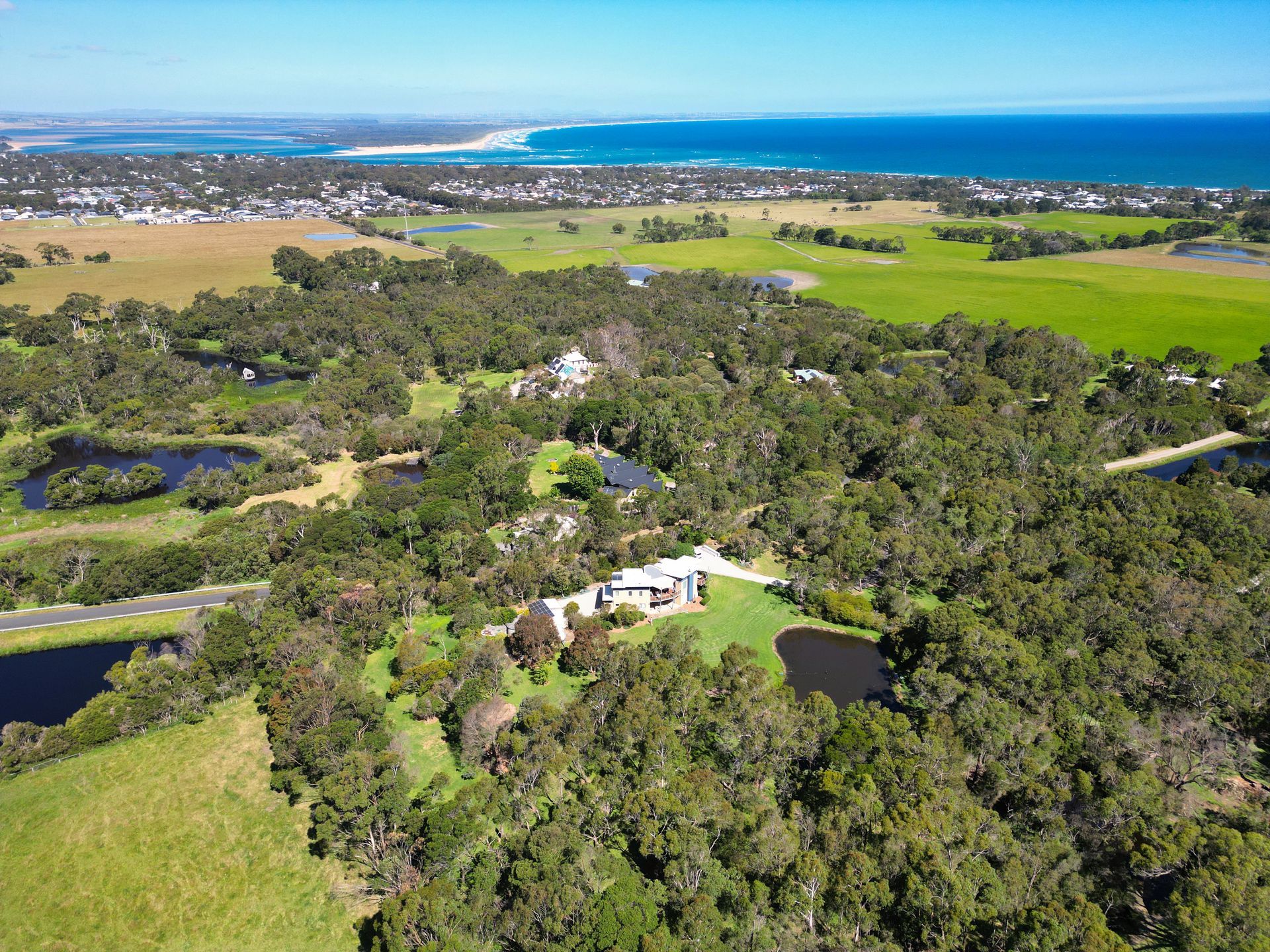 Aerial view of a house nestled in a lush green area with ponds, fields, and a coastline in the background.