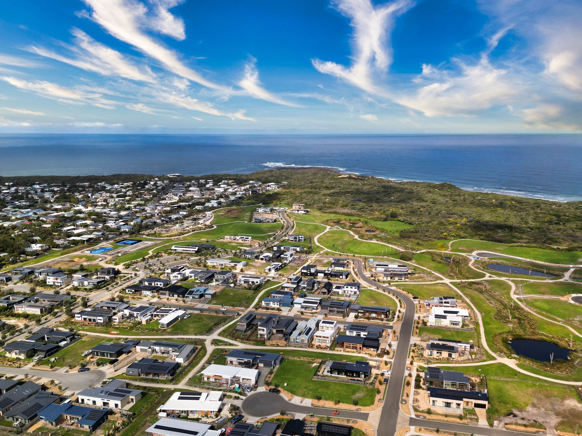 An aerial view of a modern residential neighborhood bordering a green golf course and a blue ocean under a cloudy sky.