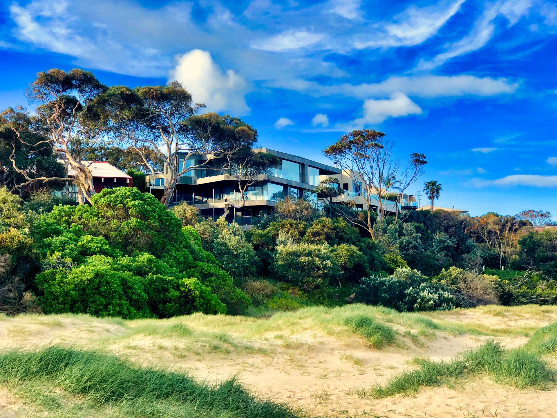 Coastal house on a dune, surrounded by green trees under a bright blue sky with clouds.