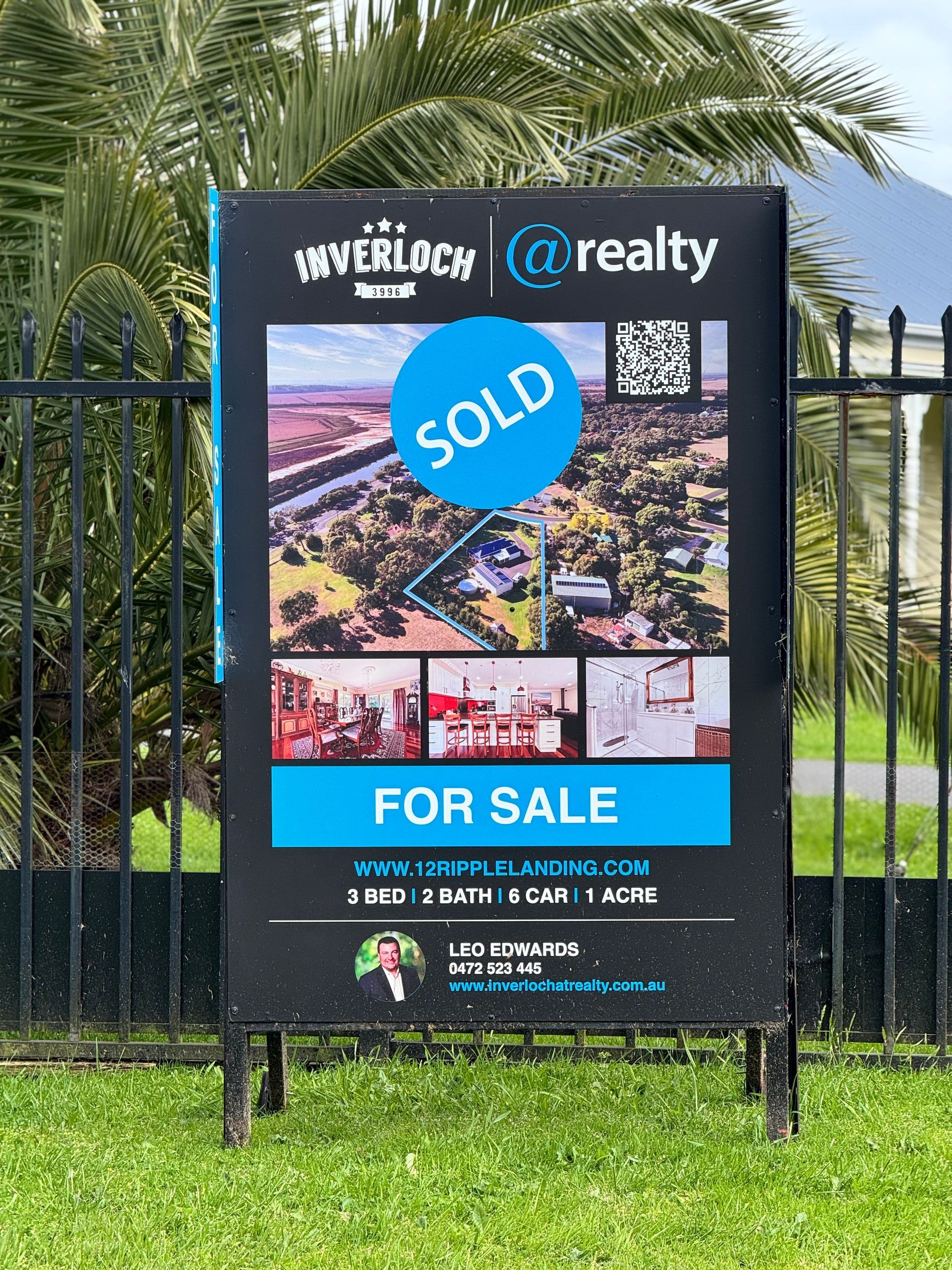 Real estate sign: SOLD property, black and blue, on grass, behind a fence.