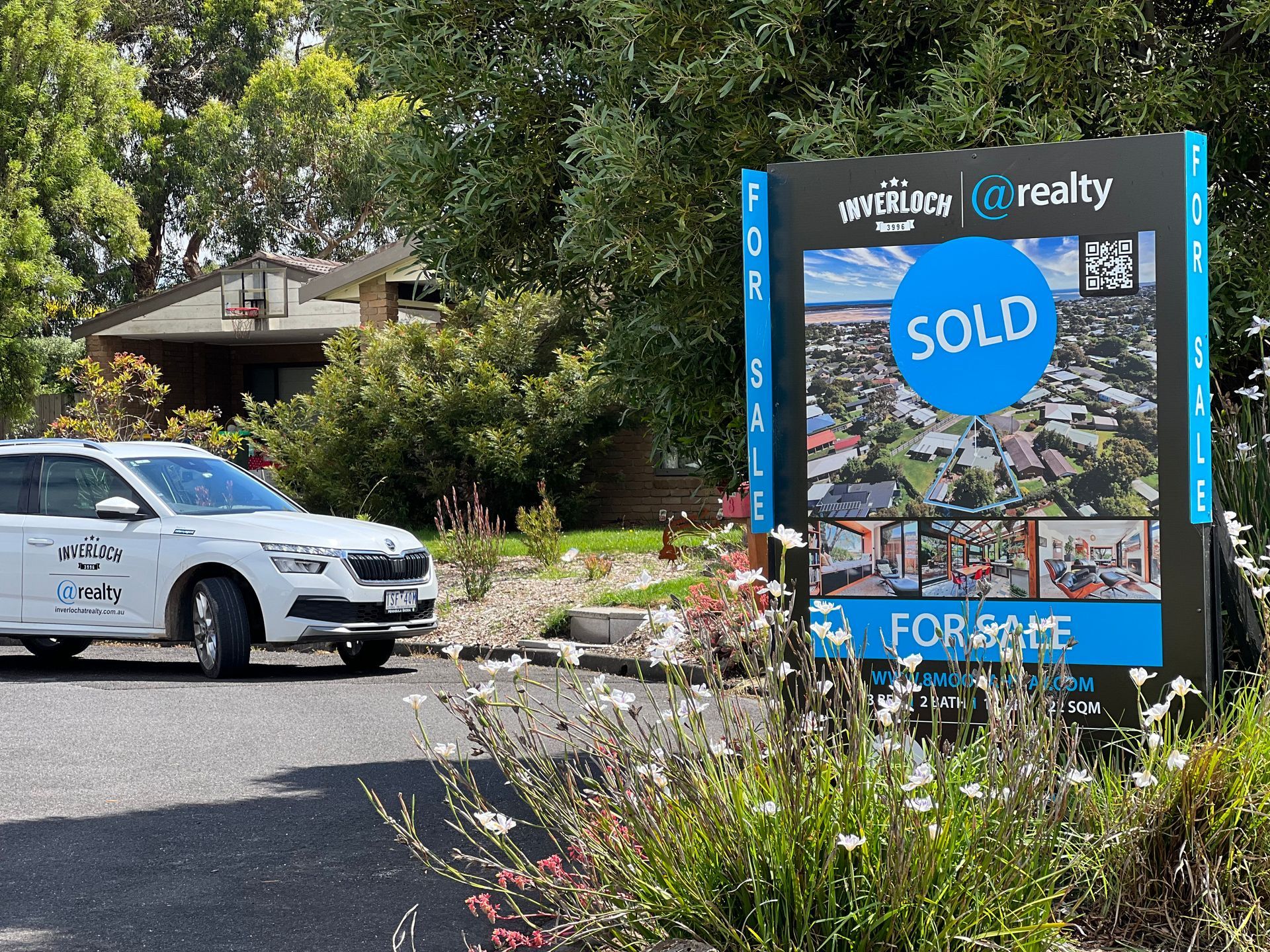 A for sold sign is surrounded by flowers and trees