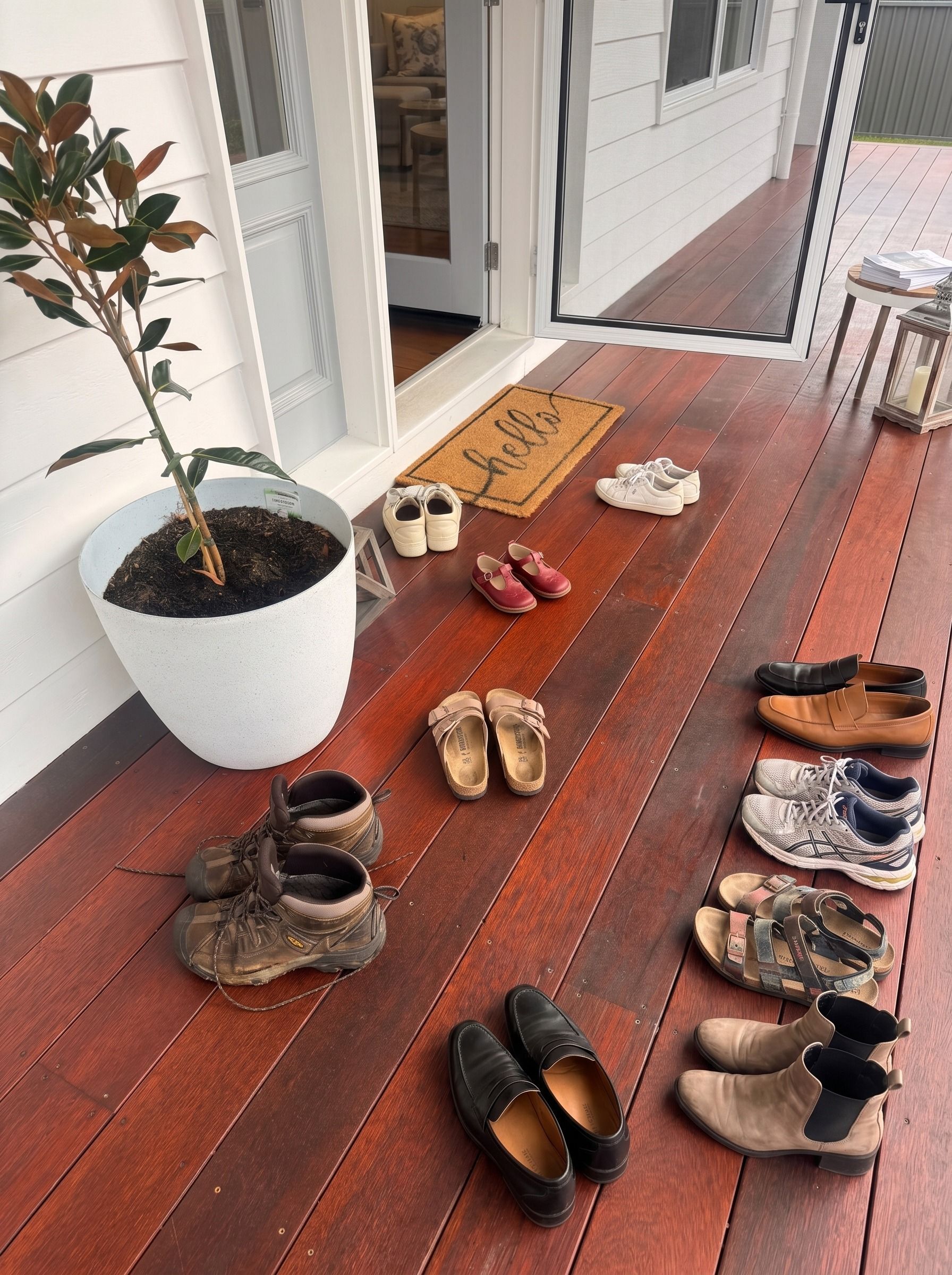 Shoes lined up on a red wooden porch beside a potted plant and welcome mat
