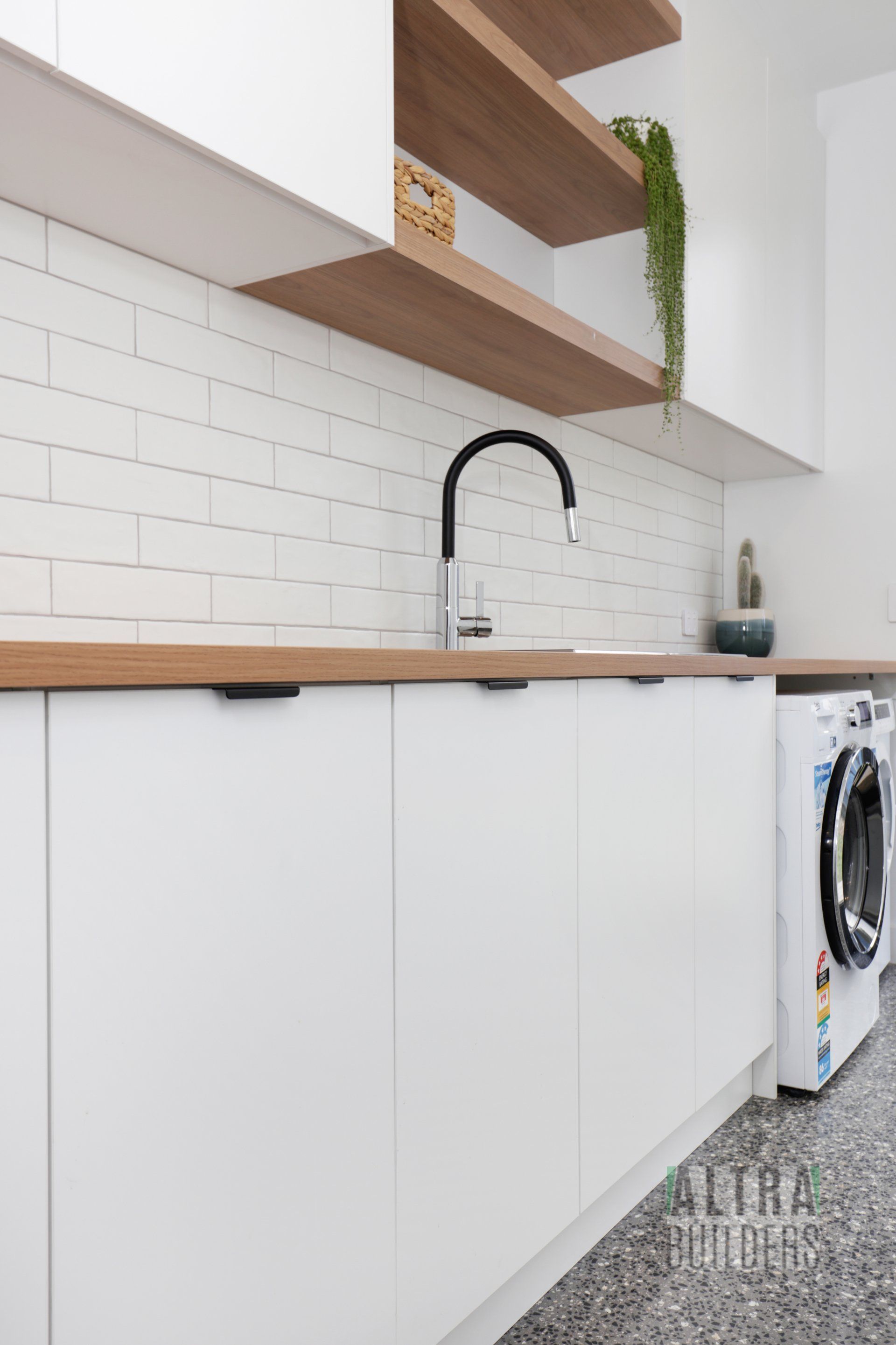 A laundry room with white cabinets , wooden shelves , a washing machine and a sink.
