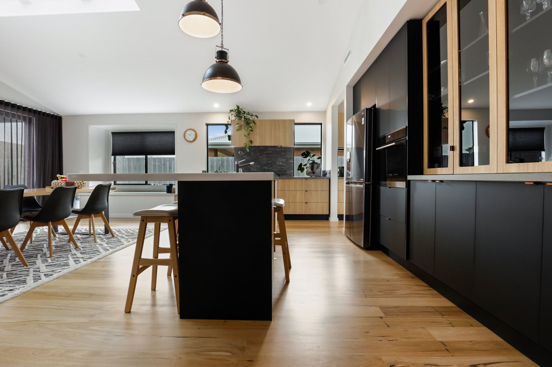 A kitchen with black cabinets and wooden floors and a dining room in the background.