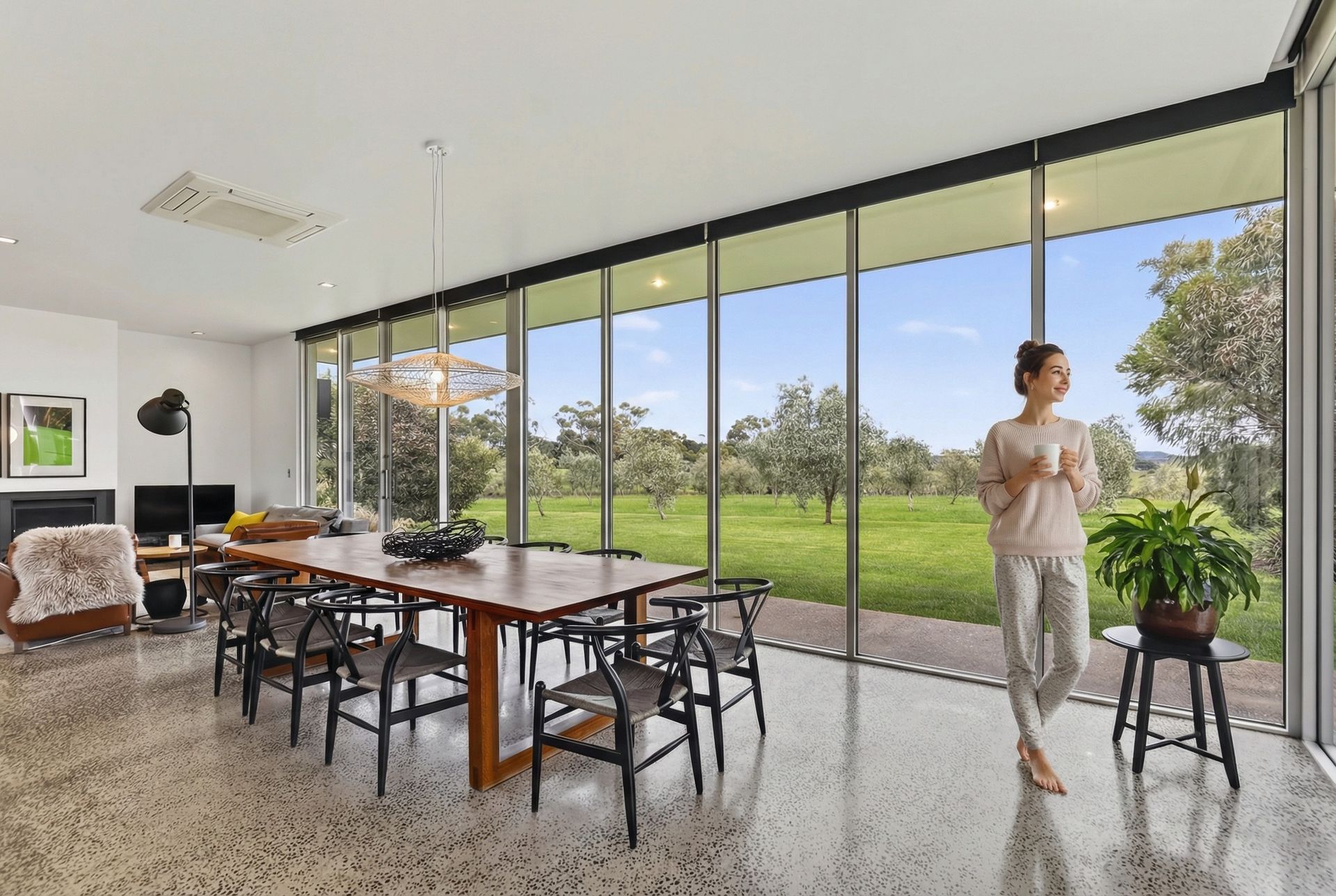 A woman with a cup stands by a large window, looking at a green landscape, in a modern dining room.