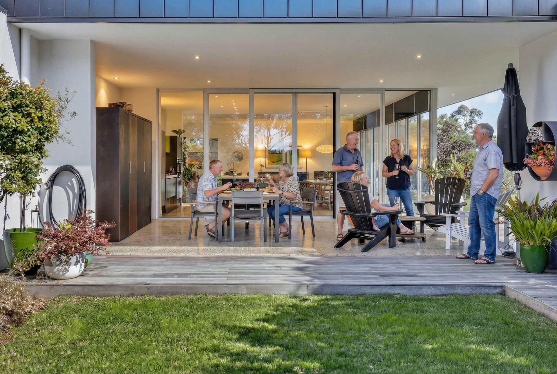 People gathered on patio, some seated at a table, others standing, near open glass doors, overlooking green lawn.
