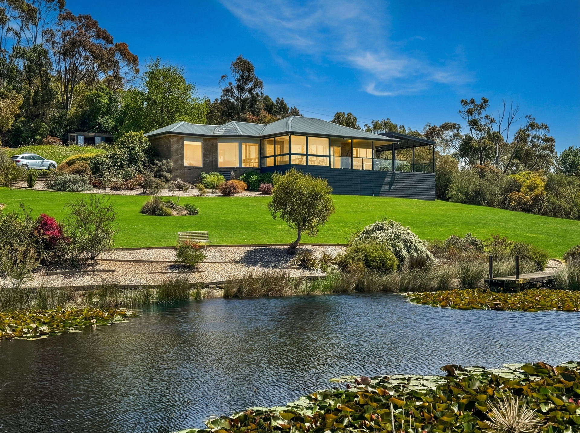 A house on a green hillside beside a pond, with trees, flowers, and a bright blue sky.