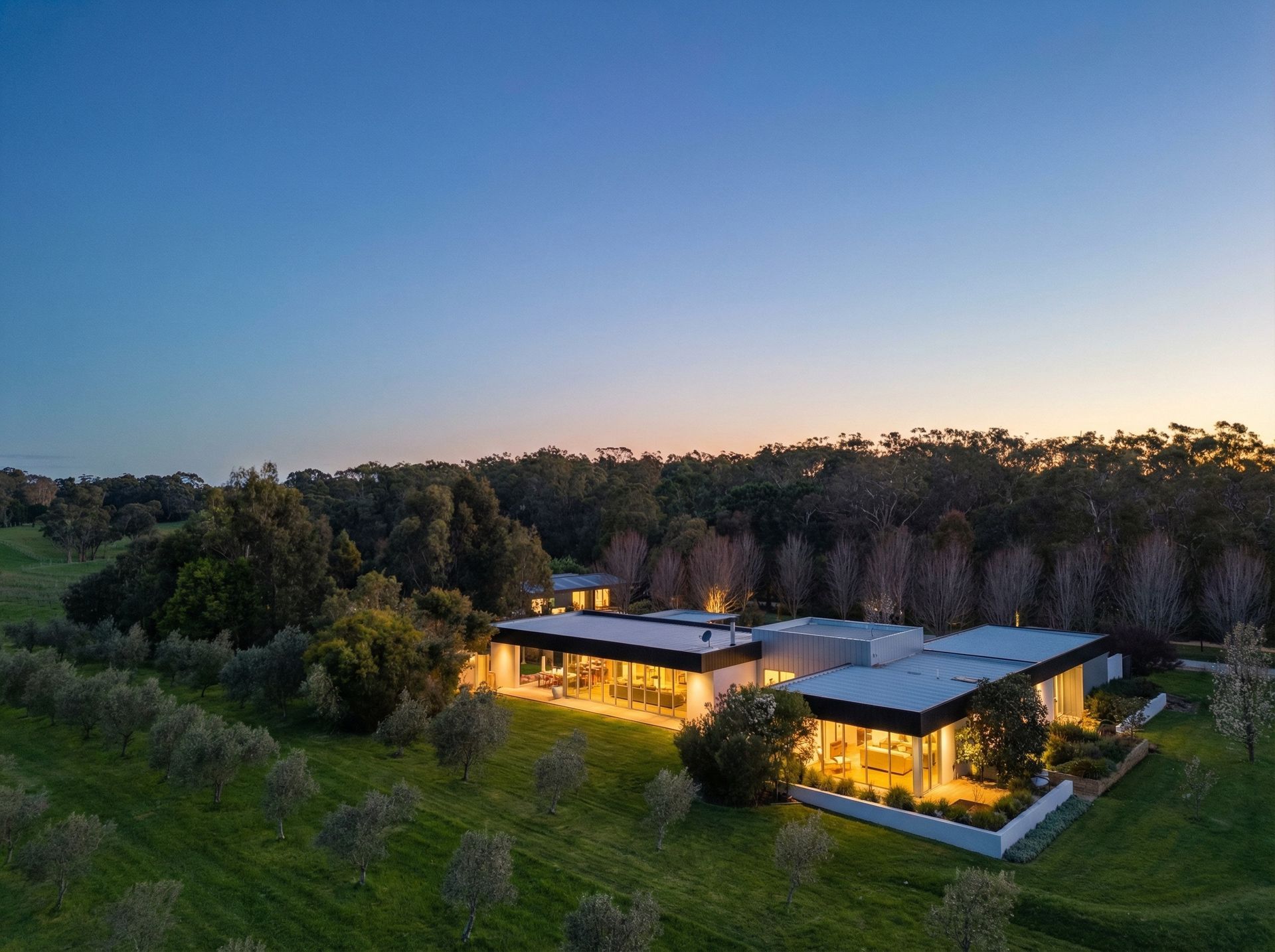 Modern home at dusk with lights on, surrounded by trees and a grassy field.