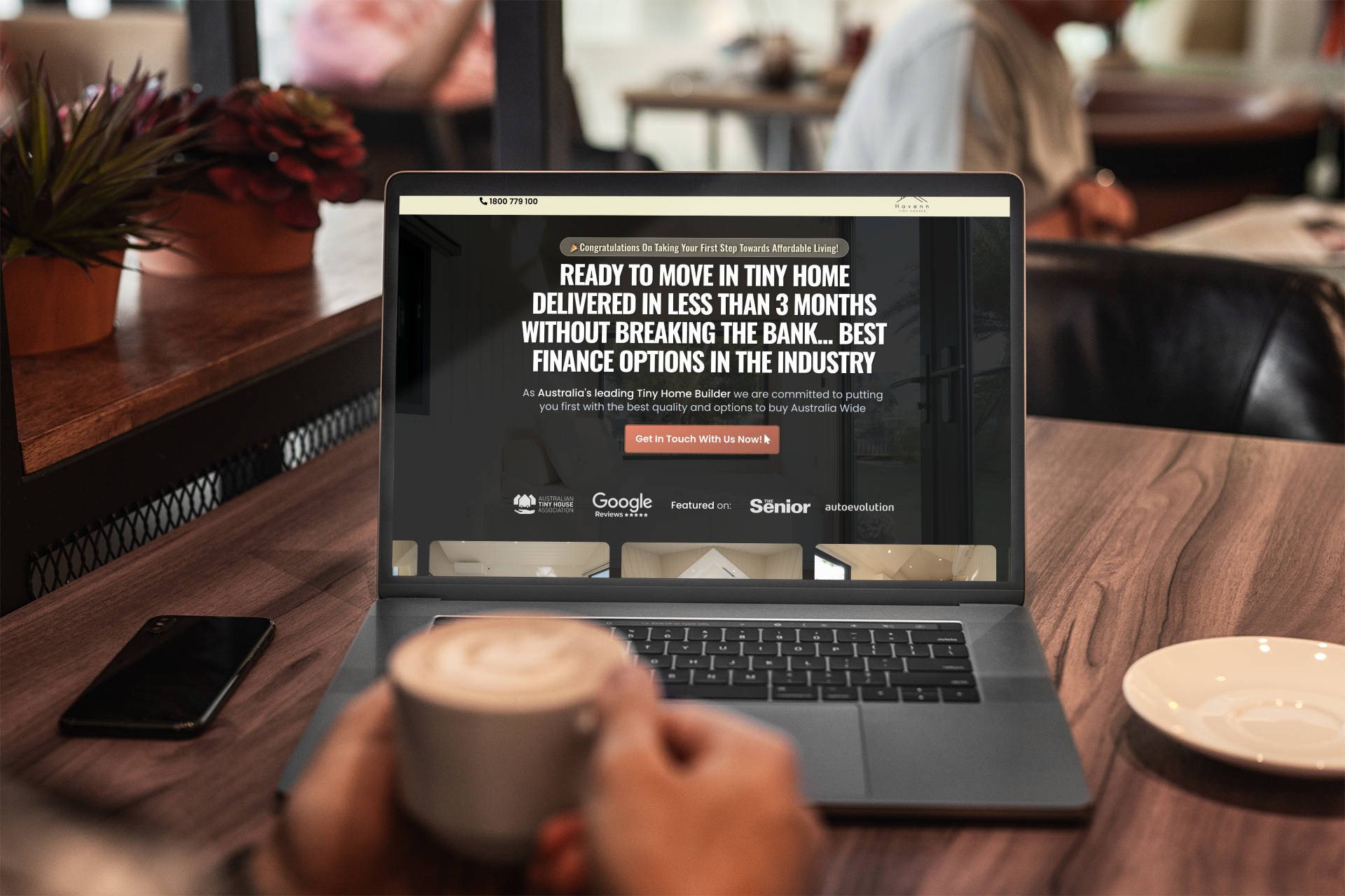 Person using a laptop with a website on a wooden table, holding a coffee cup in a cafe.