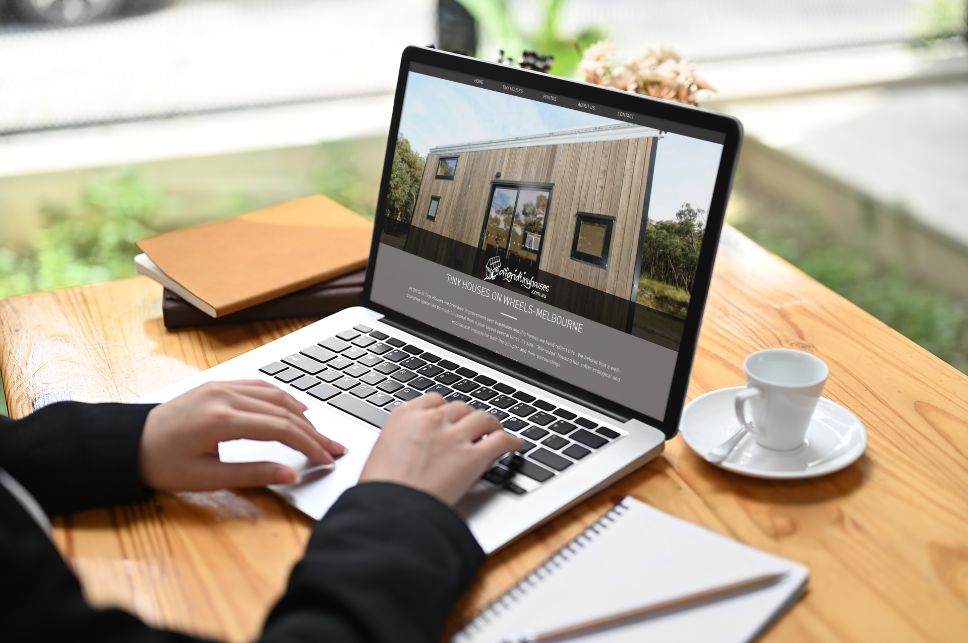 Person using laptop to browse a website featuring a tiny house design; coffee and notebook on the table.https://www.offgridtinyhouses.com.au/