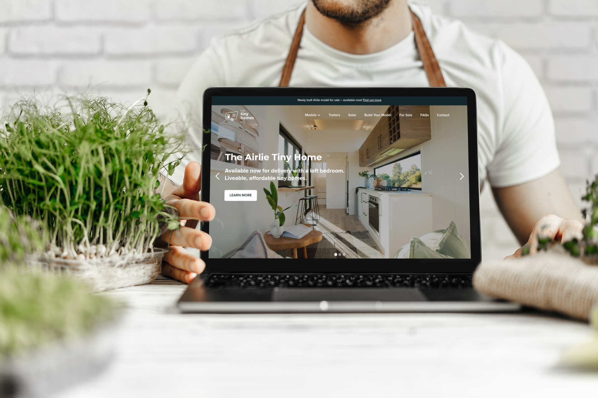 Man holding laptop displaying a website for interior design, with greenery nearby.