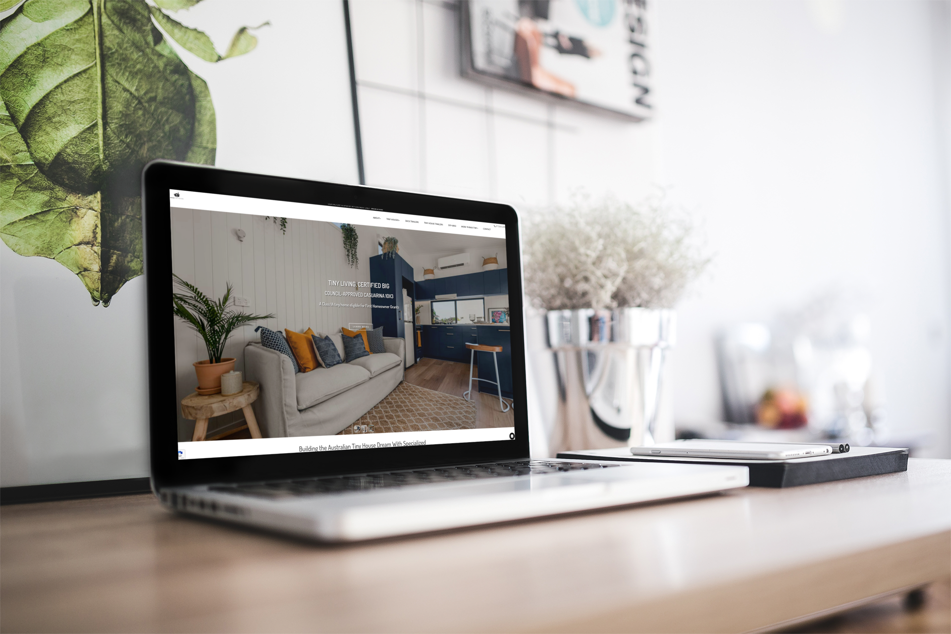 Laptop displaying a website about interior design, on a desk with flowers and a framed print.