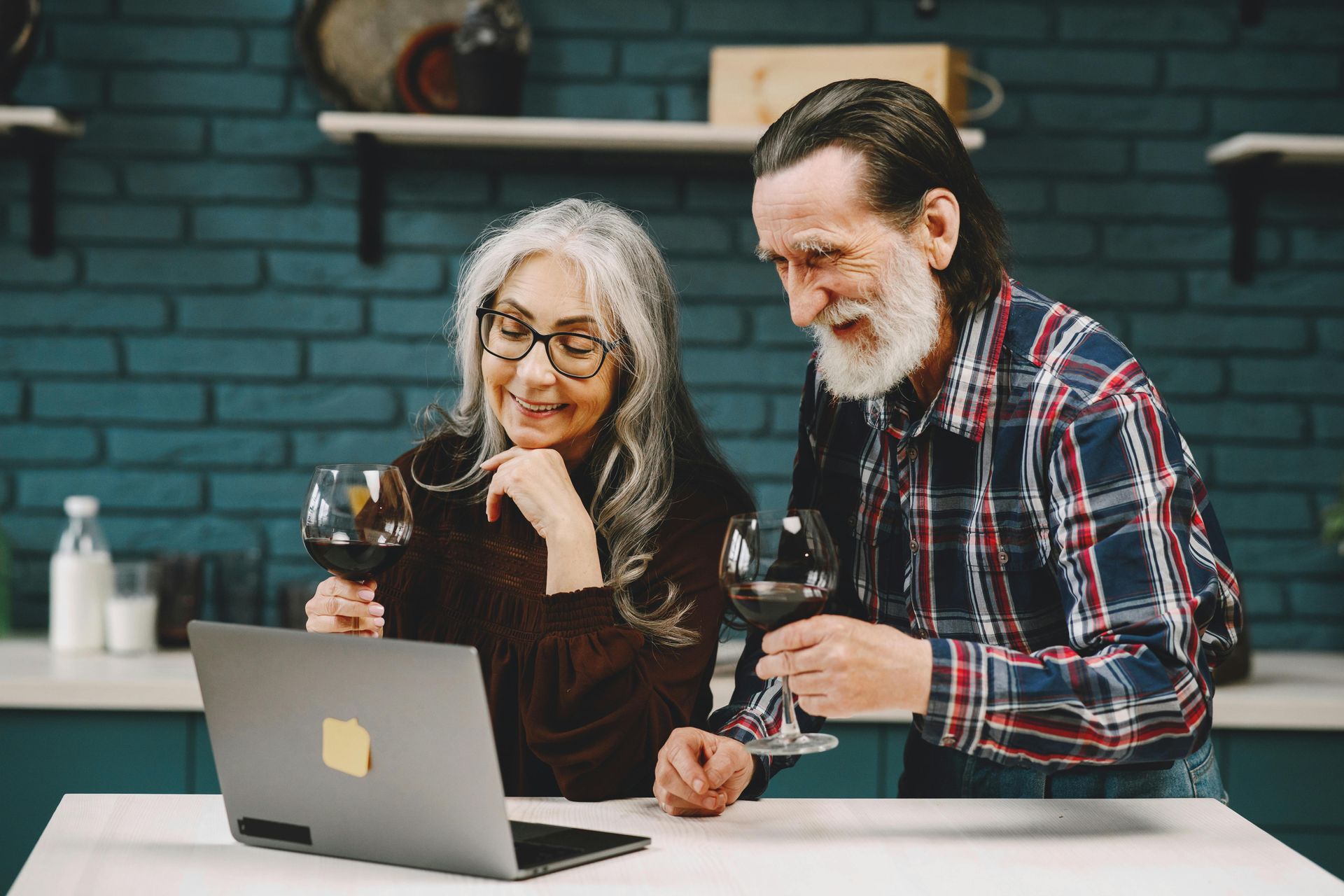 Couple toasting with wine, looking at a laptop in a kitchen.