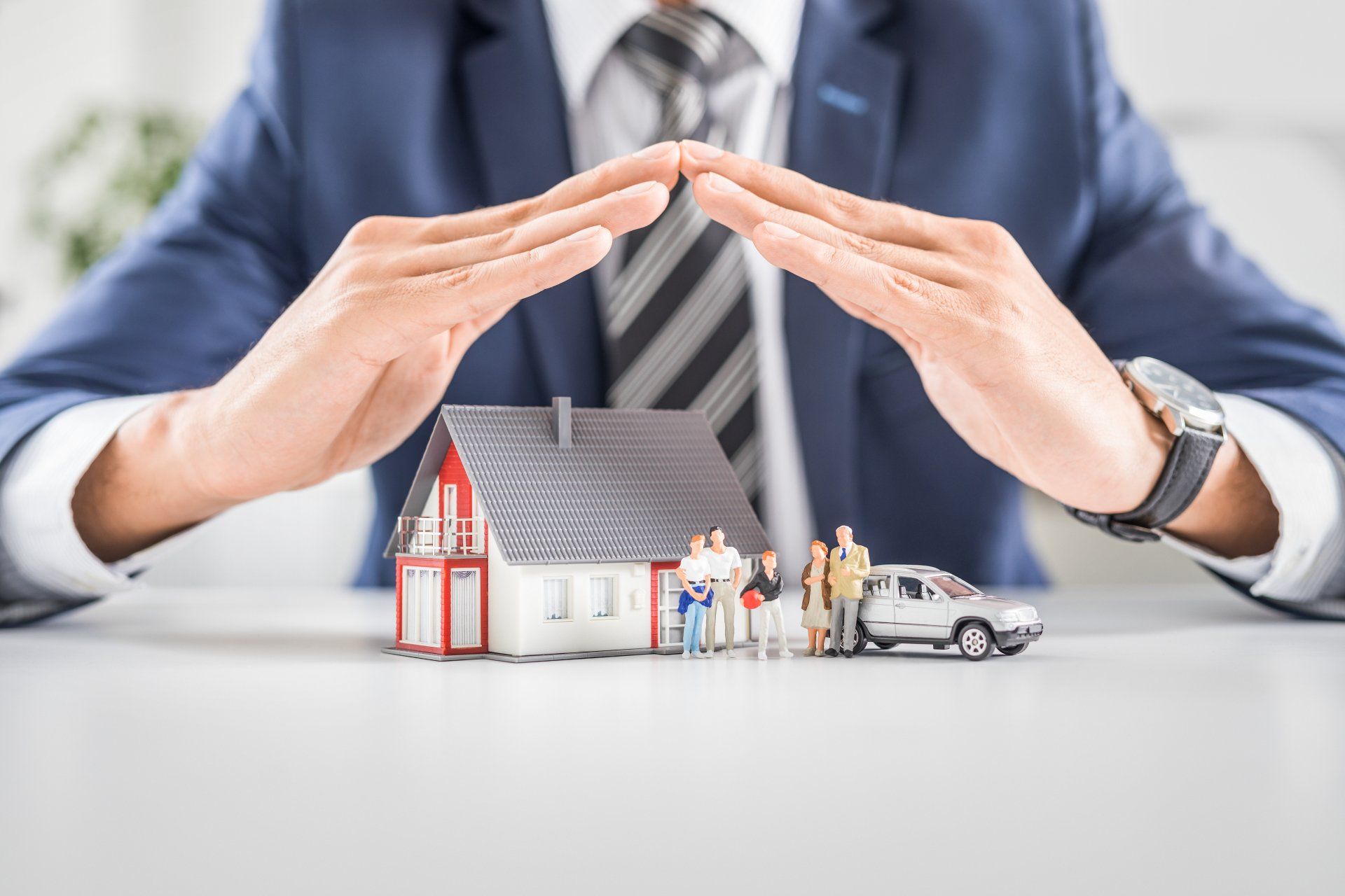 A man in a suit and tie is sitting at a table covering a house and a car with his hands.
