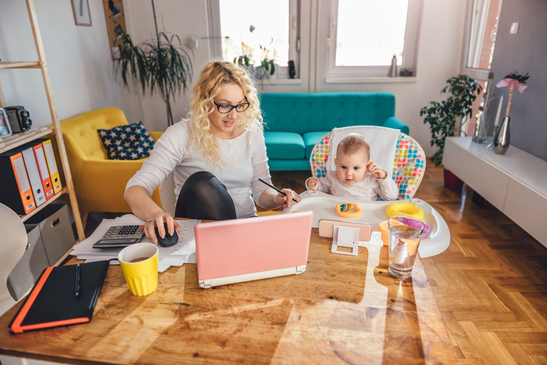 A woman is sitting at a desk with a laptop and a baby.