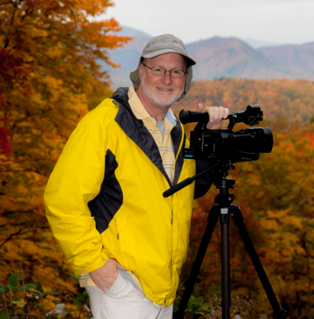 Groom kissing bride's forehead under a veil. They embrace, smiling; sunlit outdoor setting.