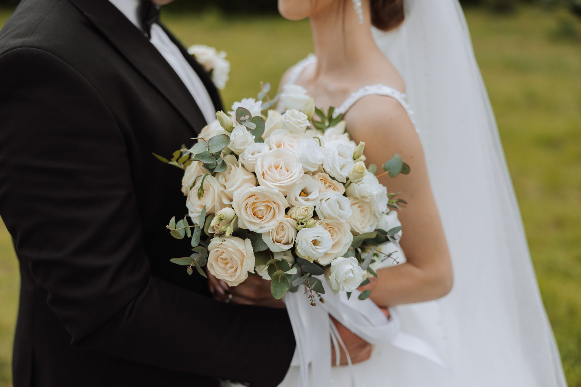 Bride and groom embrace, bride holding a bouquet of cream-colored roses and greenery, outdoors.
