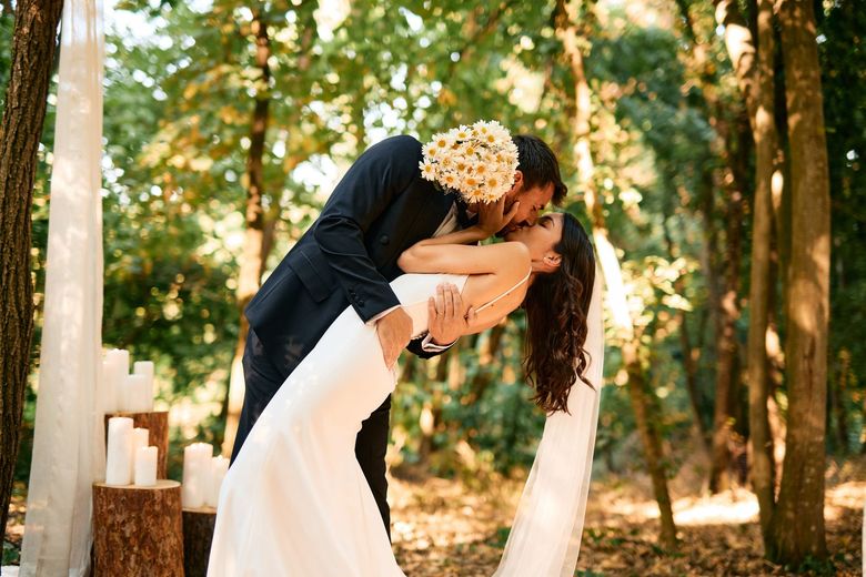 Couple kissing during outdoor wedding ceremony in a forest, bride in white dress, groom in navy suit.