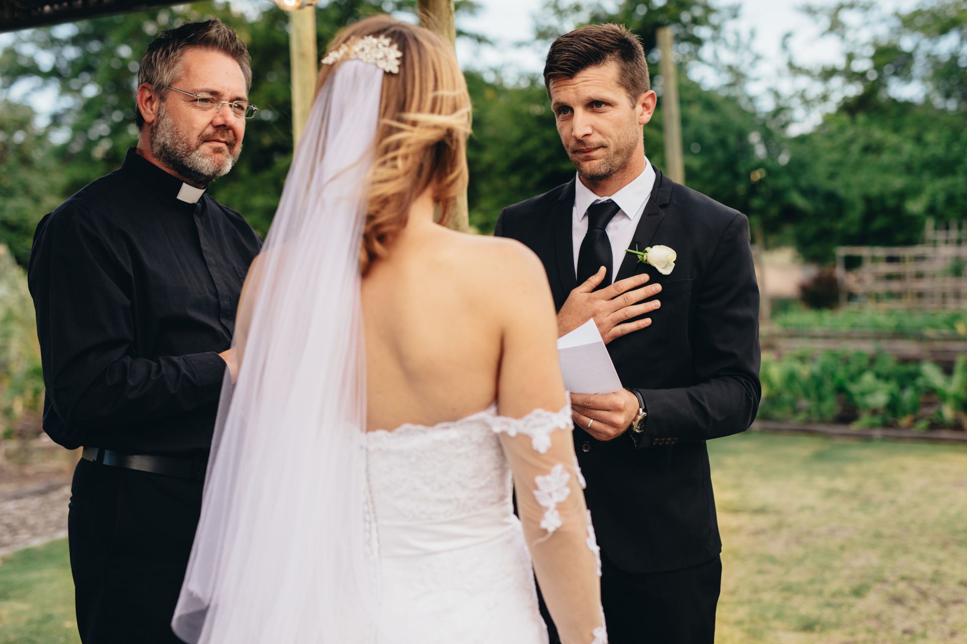 Wedding ceremony: Bride and groom facing officiant, groom with hand over heart, outdoors.