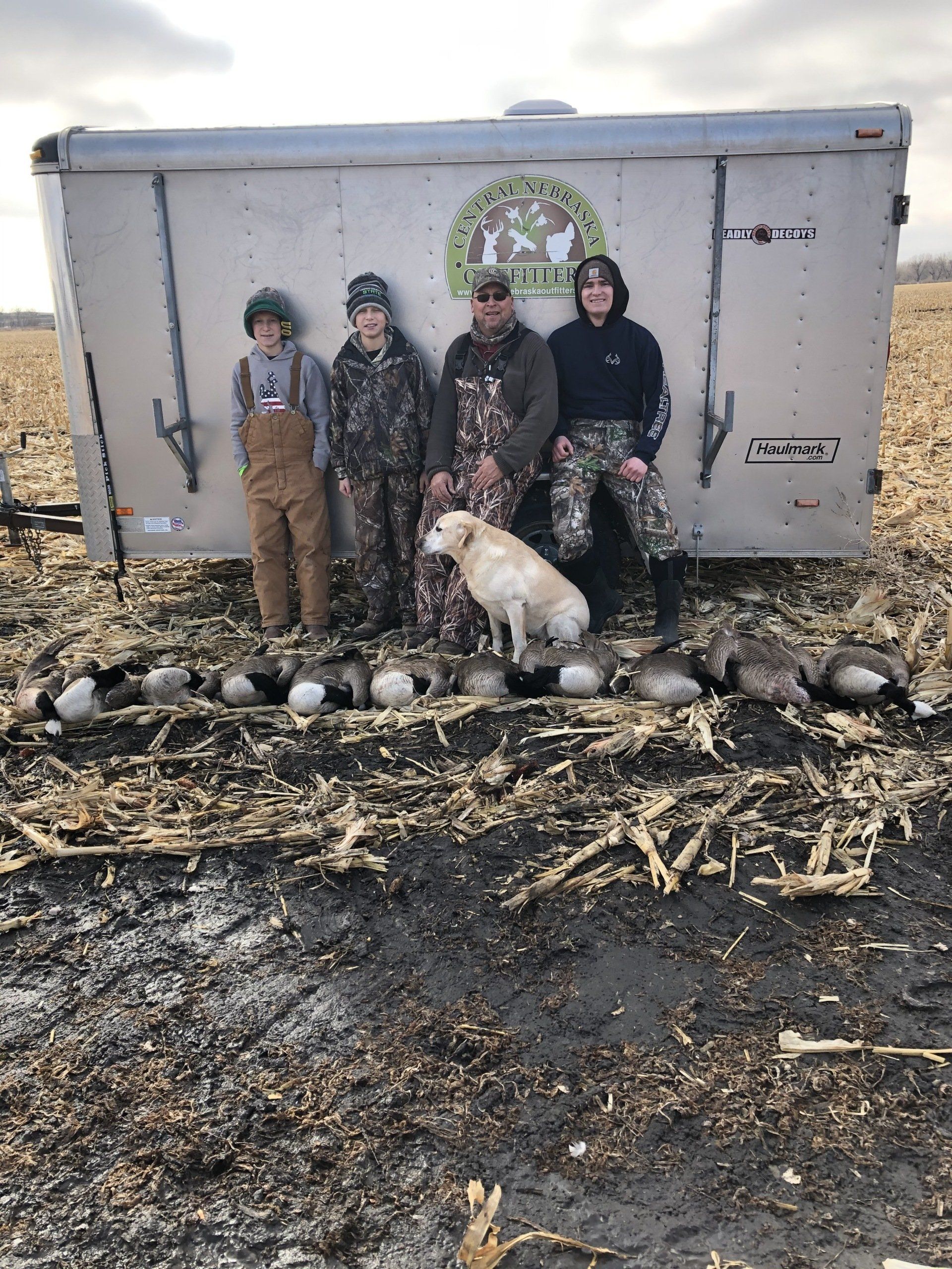 A group of people and a dog are standing in front of a trailer in a field.