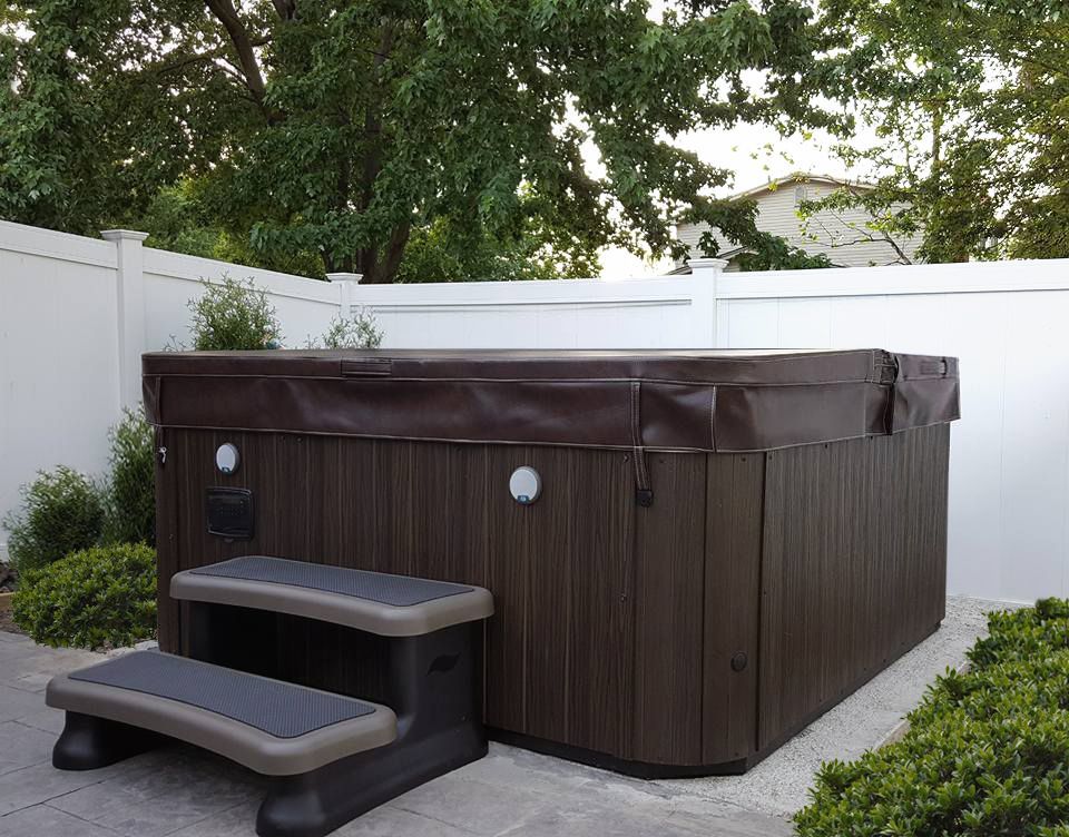 A dark brown hot tub with steps sits near a white fence, surrounded by greenery.
