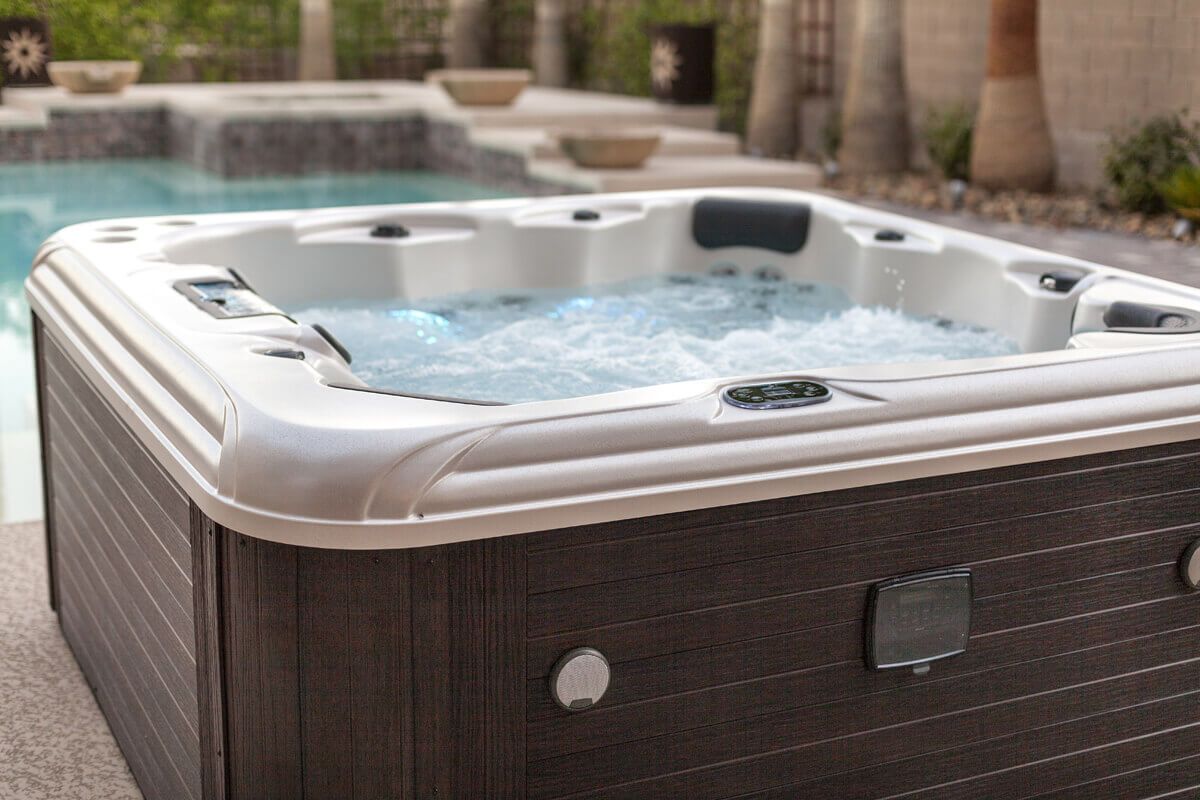 Hot tub outdoors with bubbly water, brown wicker-style siding, and a pool in the background.