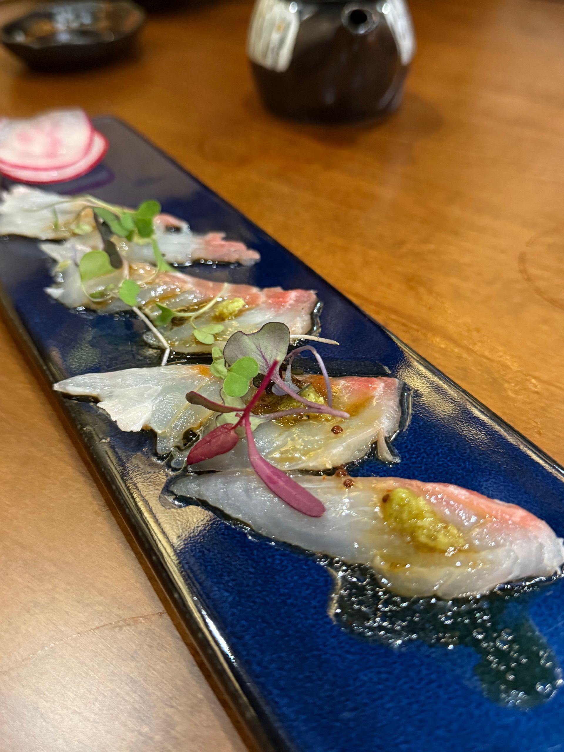 Sliced sashimi garnished with herbs on a long blue plate, served on a wooden table.