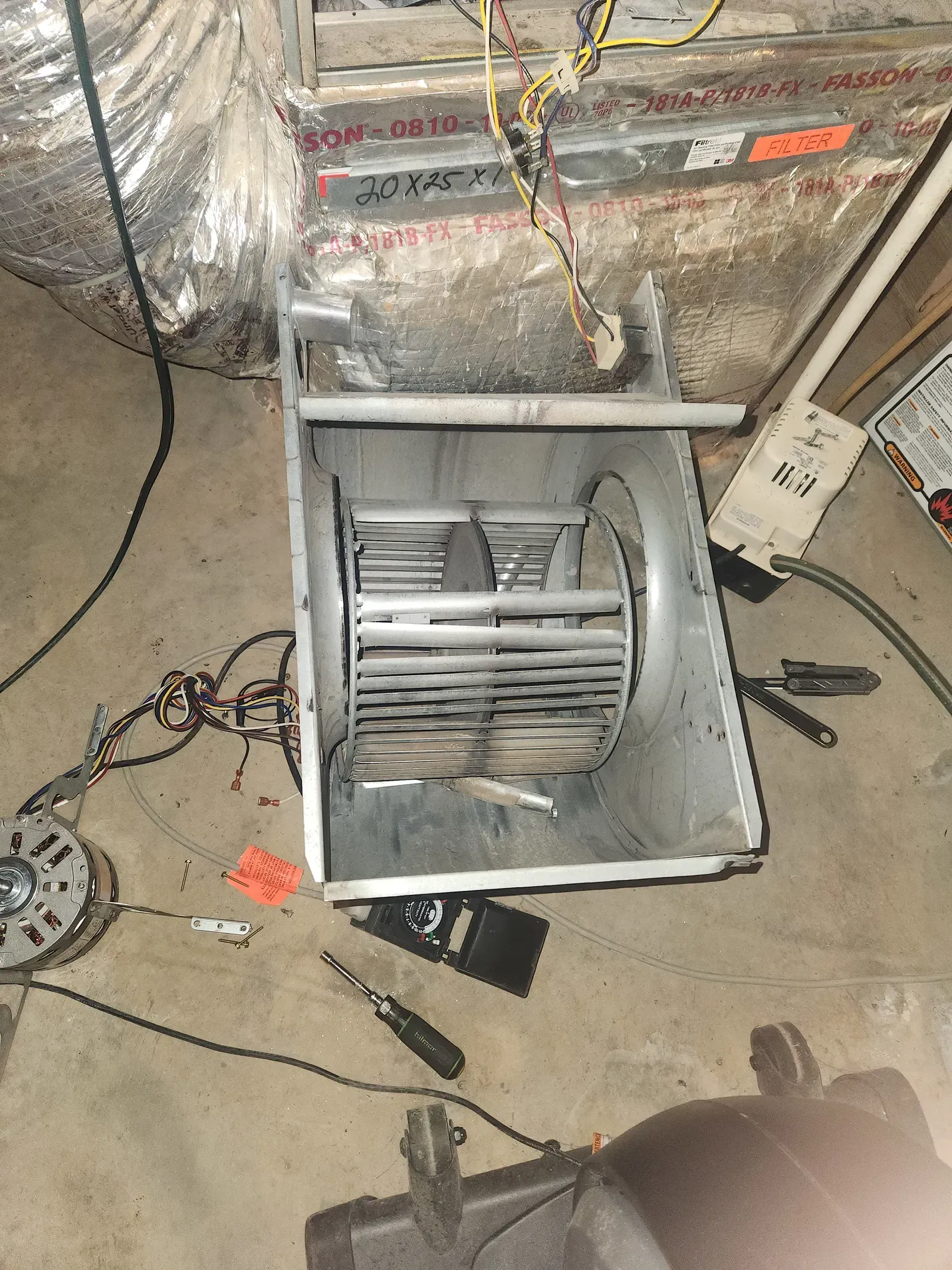 Furnace blower assembly, disassembled.  Metal fan blades inside a housing, tools on the concrete floor.