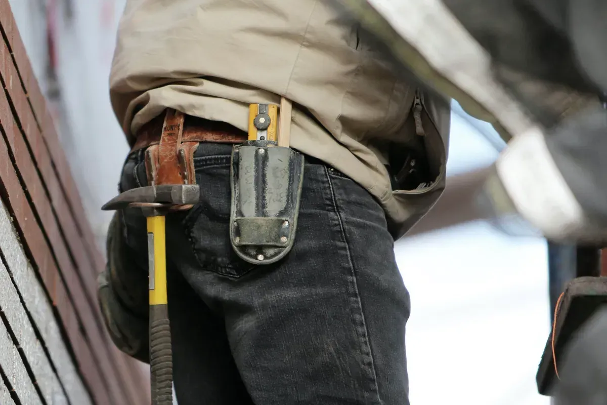 Worker with a tool belt, hammer, and knife holster. Yellow tool handle. Dark jeans, beige jacket.