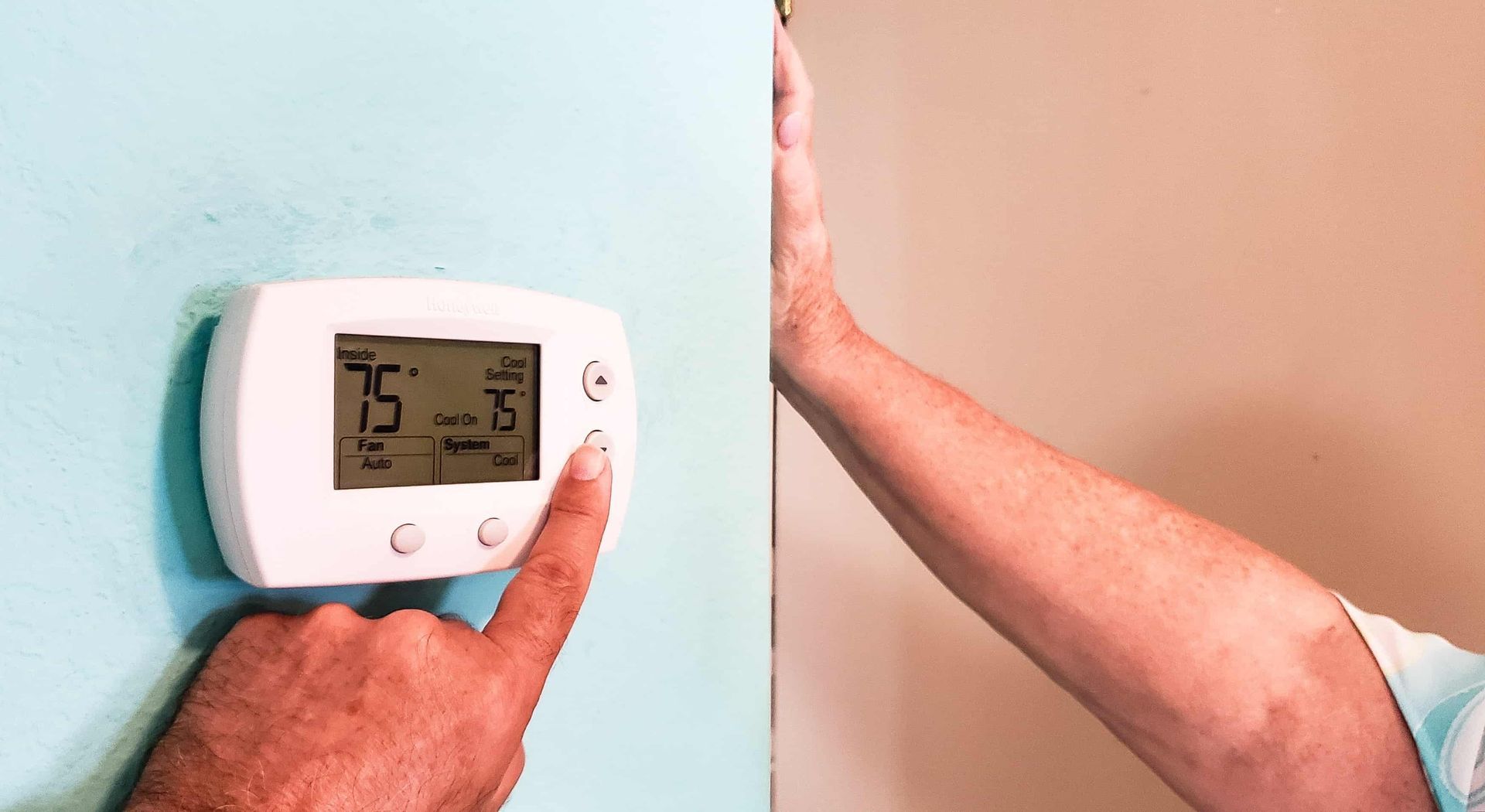 A hand adjusting a digital thermostat set to 75 degrees on a blue wall, next to a person.
